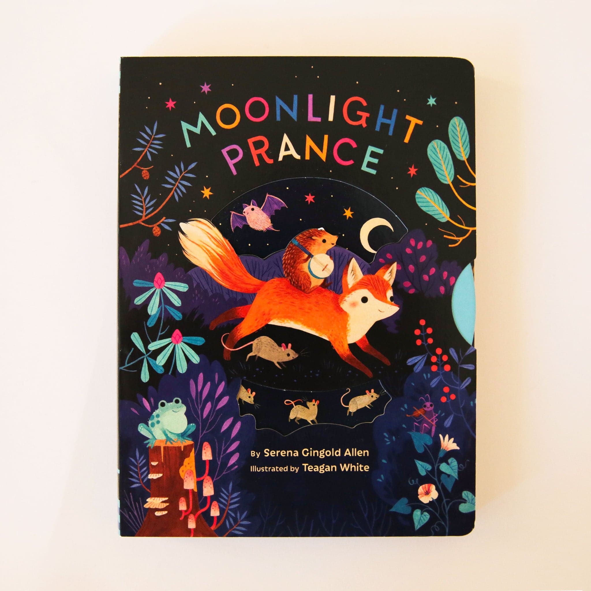 Hard cover children's book titles 'Moonlight Prance' in colorful capital lettering. Below is a woodland scene of various animals dancing around in the moonlight. Animals include a smiling fox, banjo playing hedgehog, mice and more. The background of the cover is solid black and accented with colorful star detailing. 