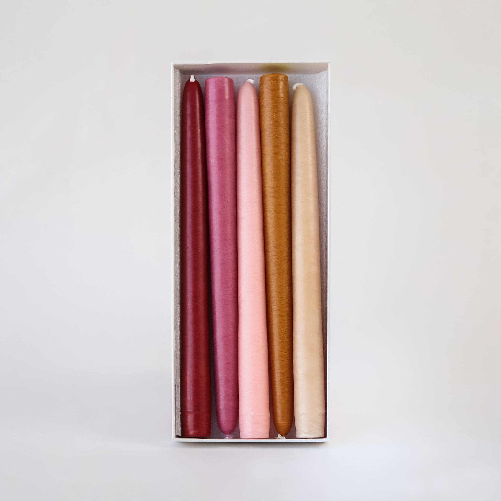 Set of five, 10 inch taper candles that come in warm toned red, pinks and oranges laying in white box against white background. 