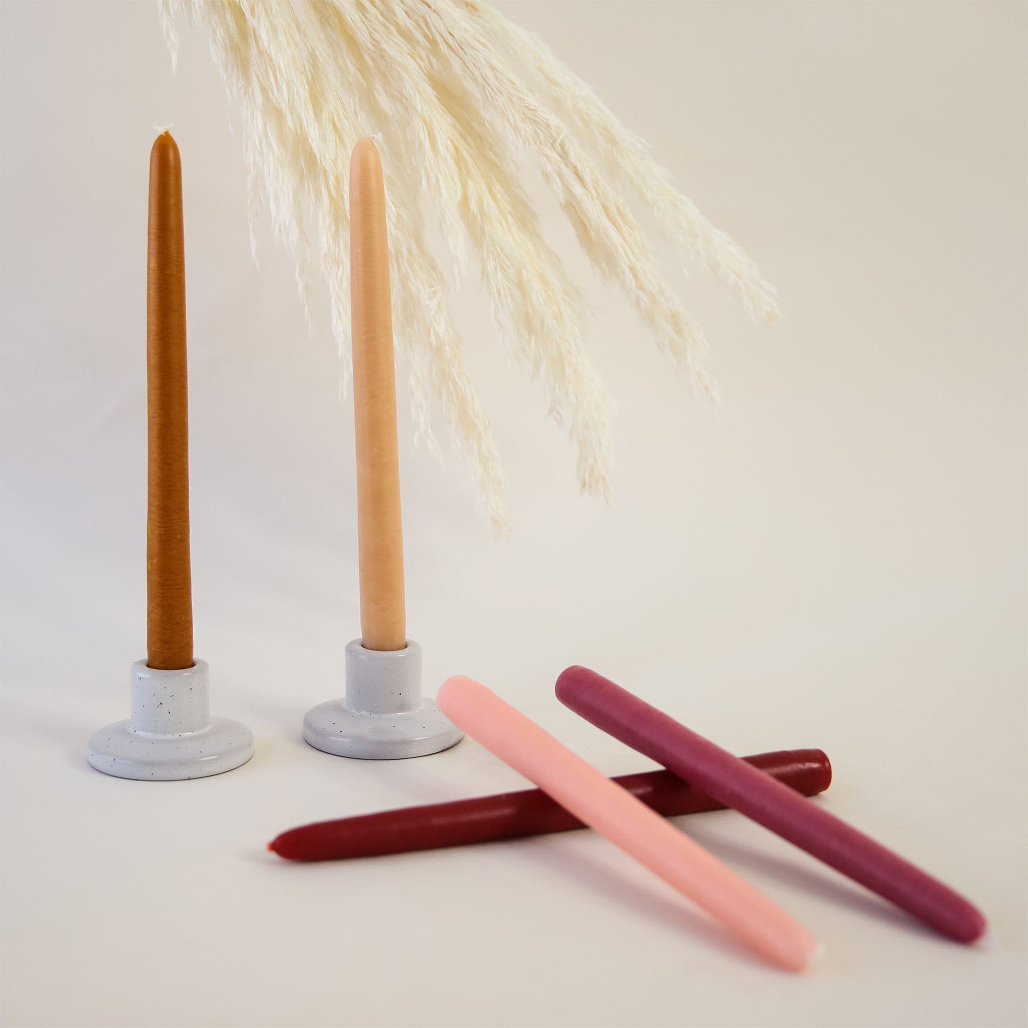 Two light and dark orange toned 10 inch candles positioned in white speckled candle holders beside three laying candles in shades of magenta. Candles are against white background with pampas grass. 