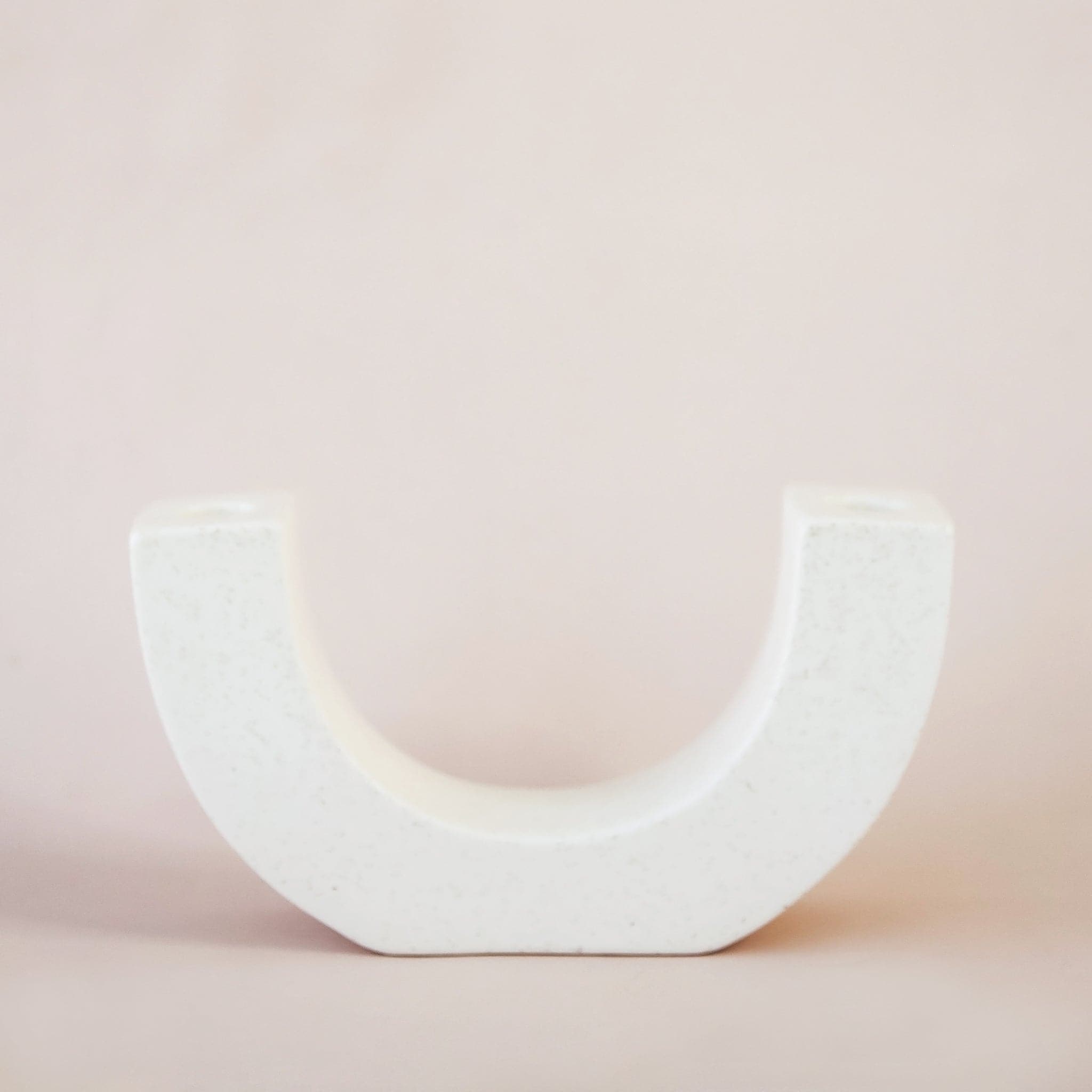 Modern ceramic u-shaped candle holder, designed to hold two candle sticks on each end. This white candle stand with faint speckles lays against a soft toned background. 