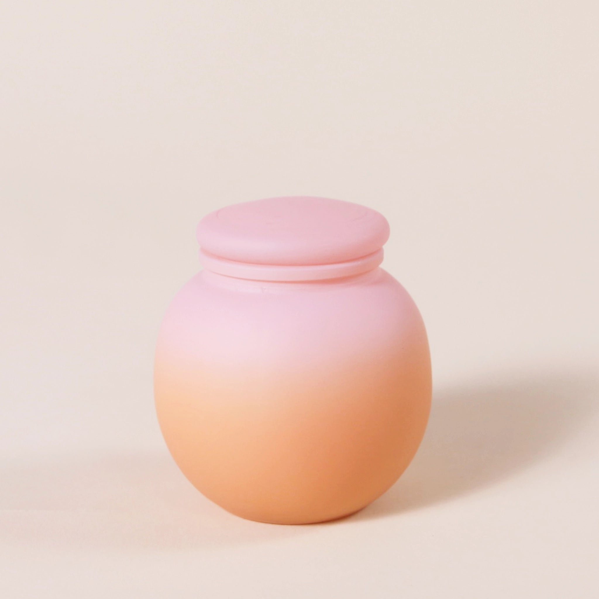 A round glass candle with a pink to orange ombre frosted finish along with a rounded lid.