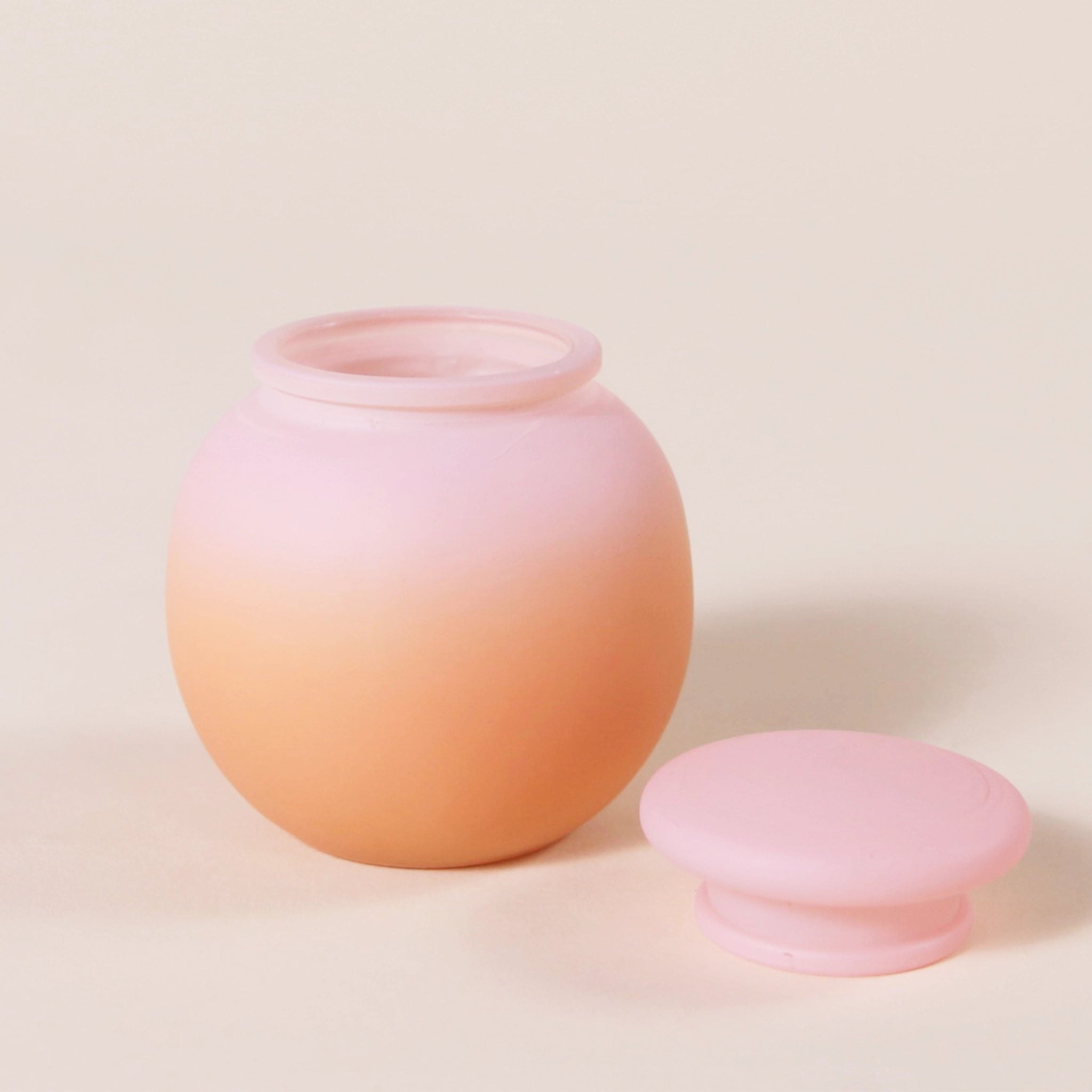 A round glass candle with a pink to orange ombre frosted finish along with a rounded lid.