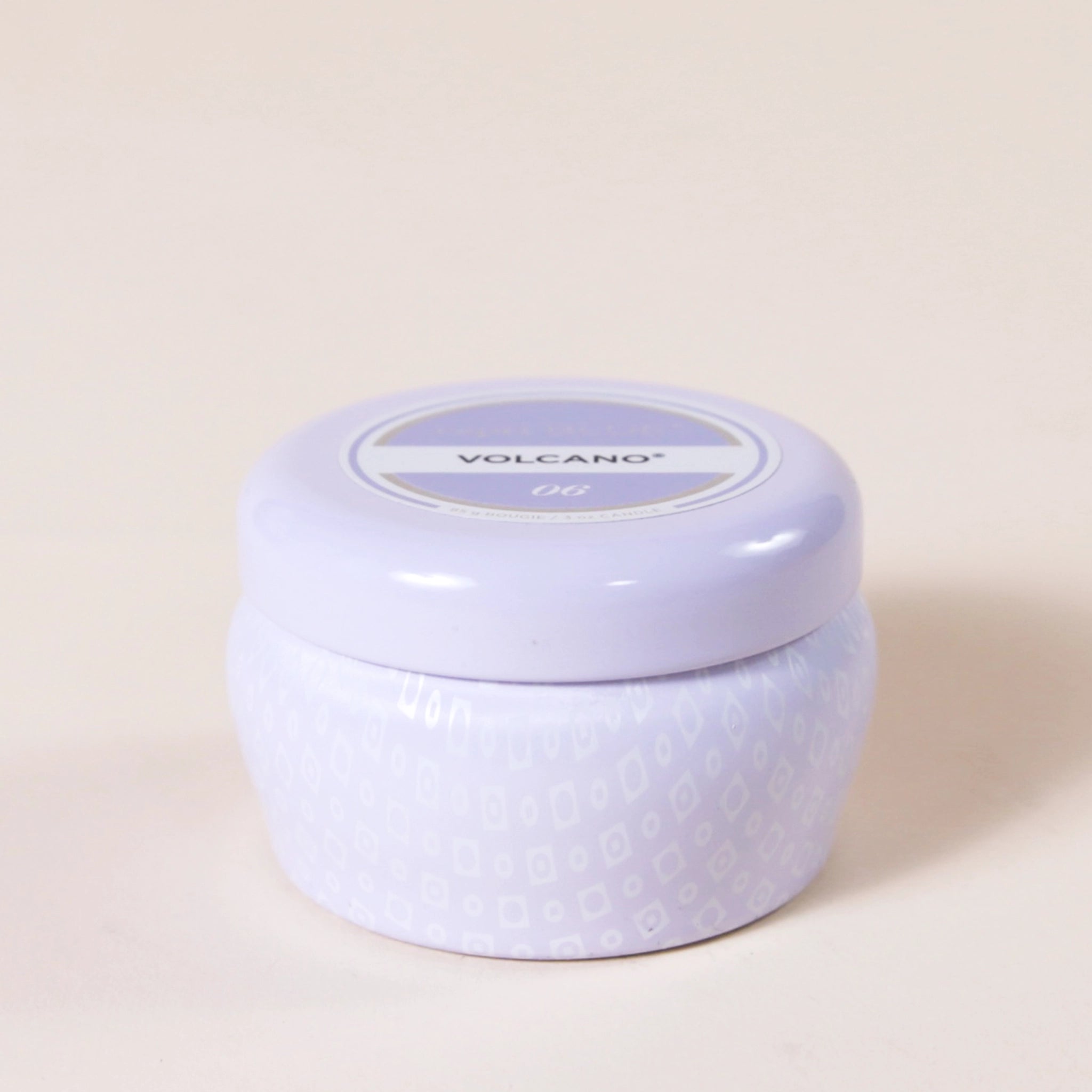 A small lavender tin candle with white wax and a white diamond repeating pattern on the outside of the tin. Comes with a matching purple lid that reads, &quot;Capri Blue Volcano&quot;.