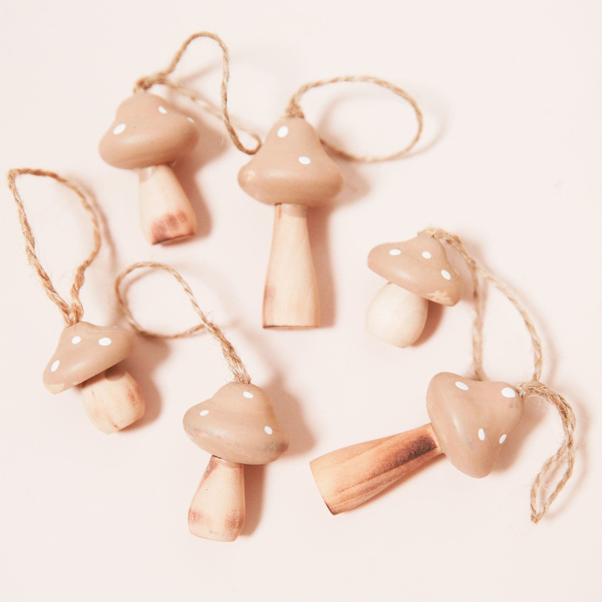 A set of 6 wooden mushroom ornaments, each a slightly differently size and shape. They have a neutral tan top with white spots and a natural wooden stem. Each ornament has an attached twine loop at the top for easy hanging. 