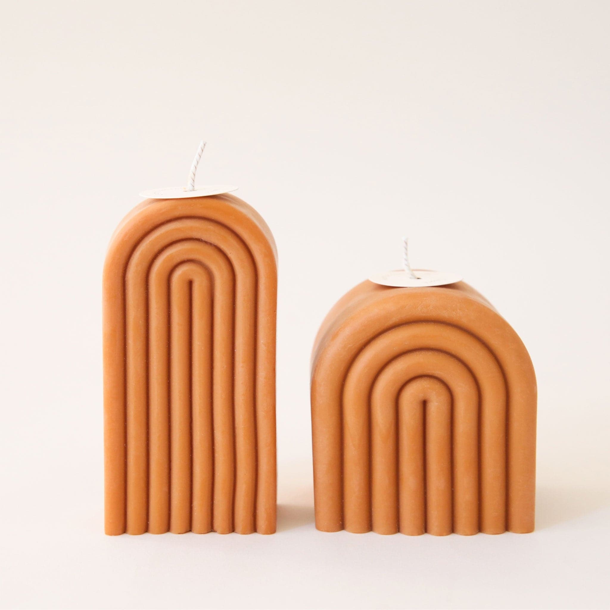 Two caramel colored arch shaped candles, one tall and one short. Each is carved with rainbow-shaped curves and has its own white wick sprouting from the top. 