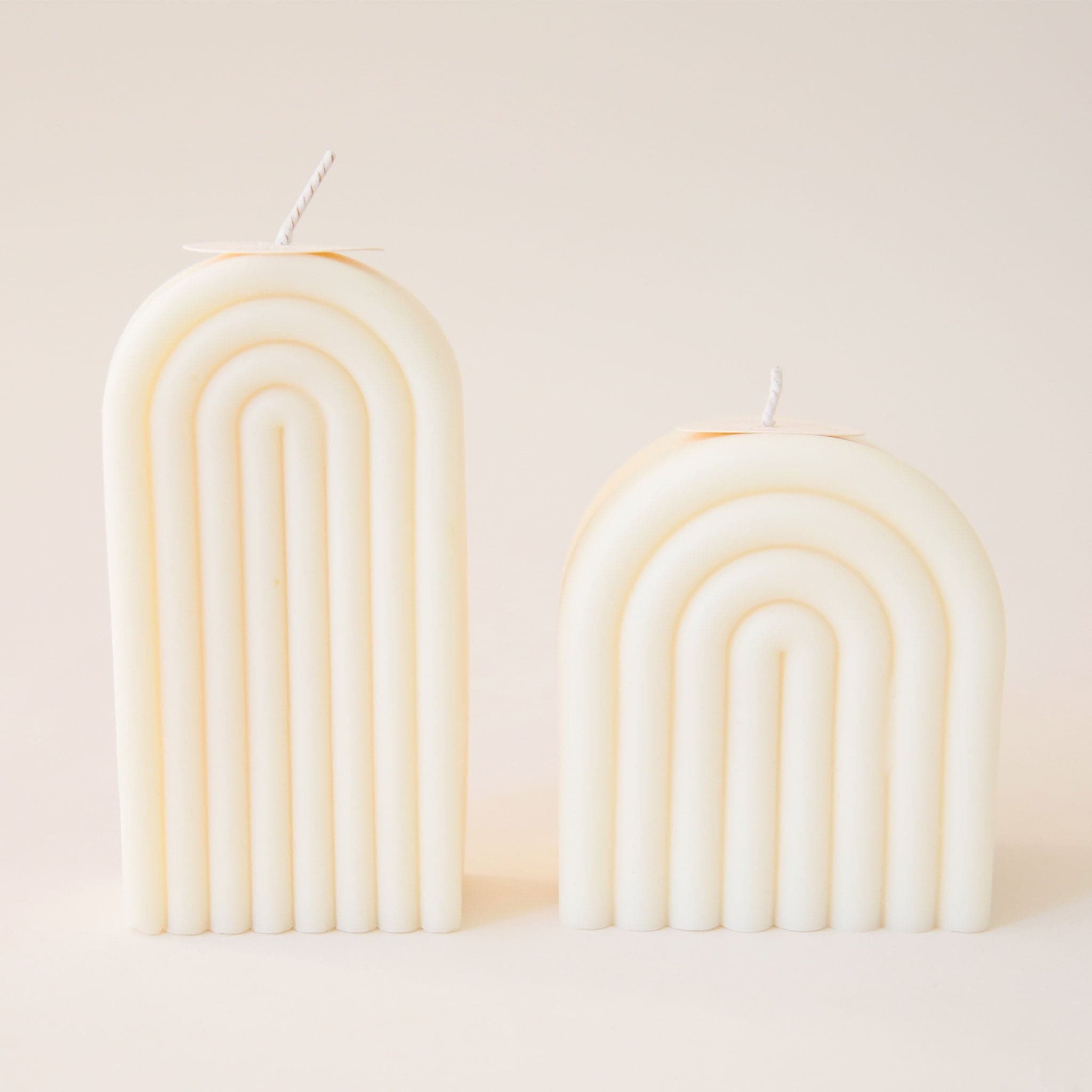 Two light cream arch shaped candles, one tall and one short. Each is carved with rainbow-shaped curves and has its own white wick sprouting from the top.
