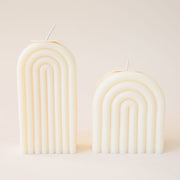 Two light cream arch shaped candles, one tall and one short. Each is carved with rainbow-shaped curves and has its own white wick sprouting from the top.