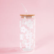 On a light pink background is a clear glass tumbler with a wood lid, a glass straw and white daisy designs all over.