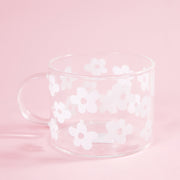 On a light pink background is a clear glass mug with a white daisy design all over.