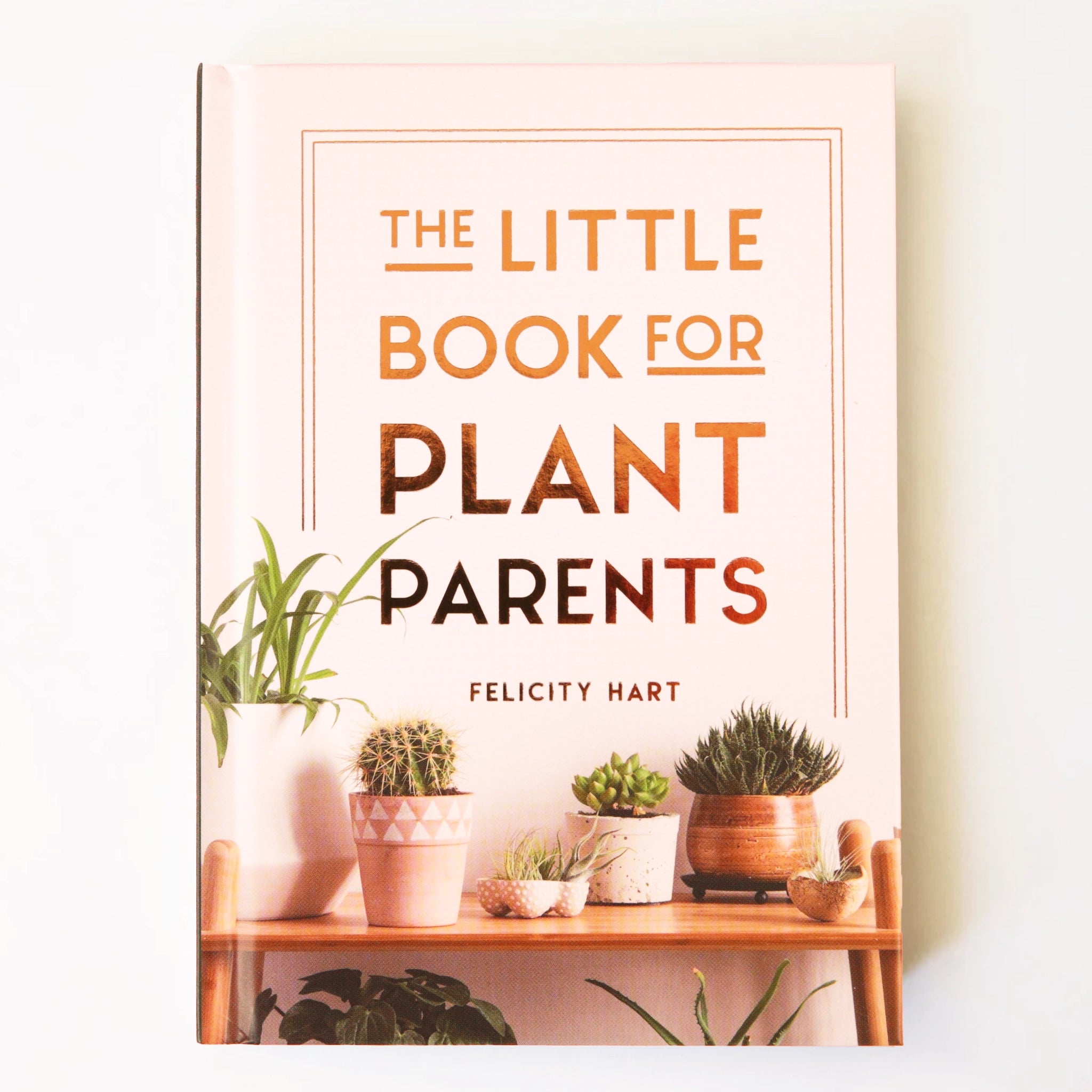 On a cream background is a light tan/pink book cover with rose gold text that reads, "The Little Book For Plant Parents by Felicity Hart" as well as a photograph of different house plants and cacti sitting on a shelf.