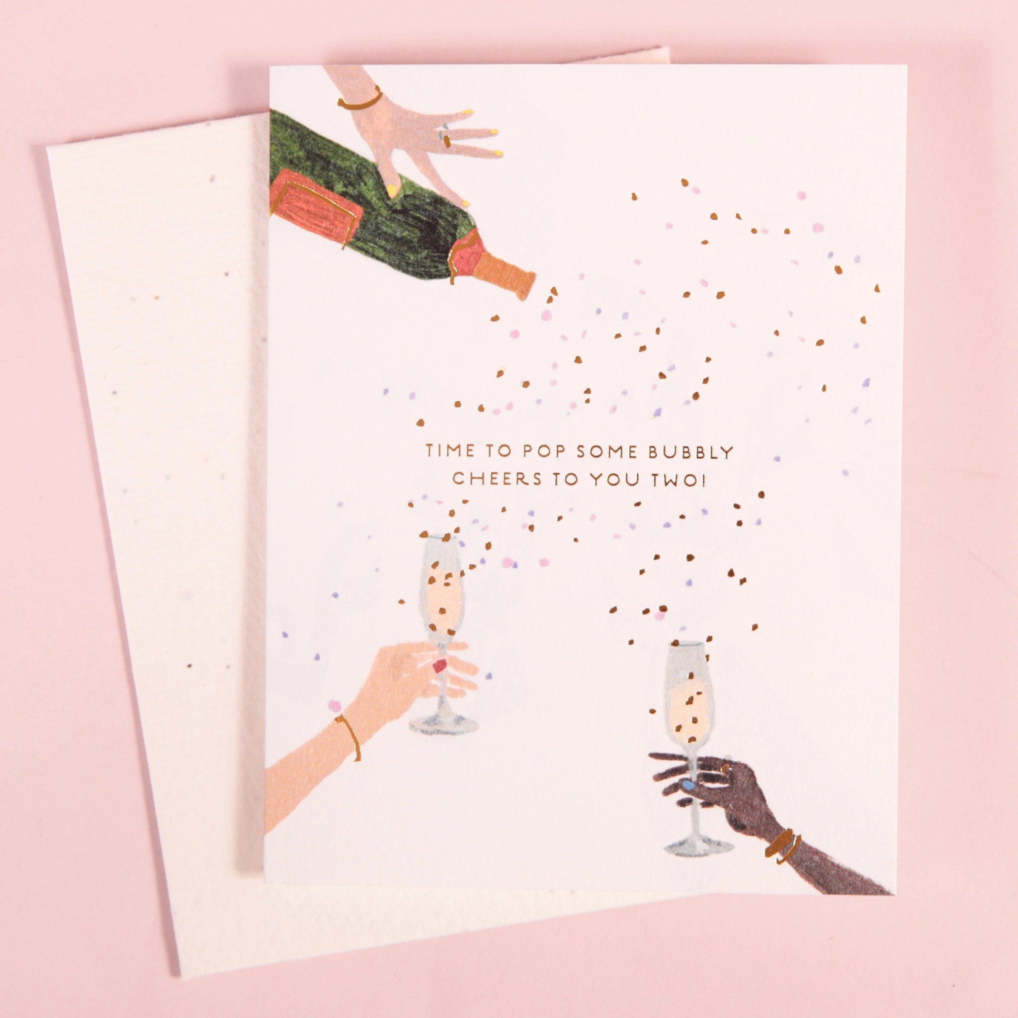 On a pink background is a white card and envelope with an illustration of three hands holding a bottle of Champagne and the others holding champagne flutes with confetti all over and small text that reads, &quot;Time to pop some bubbly cheers to you two!&quot;.