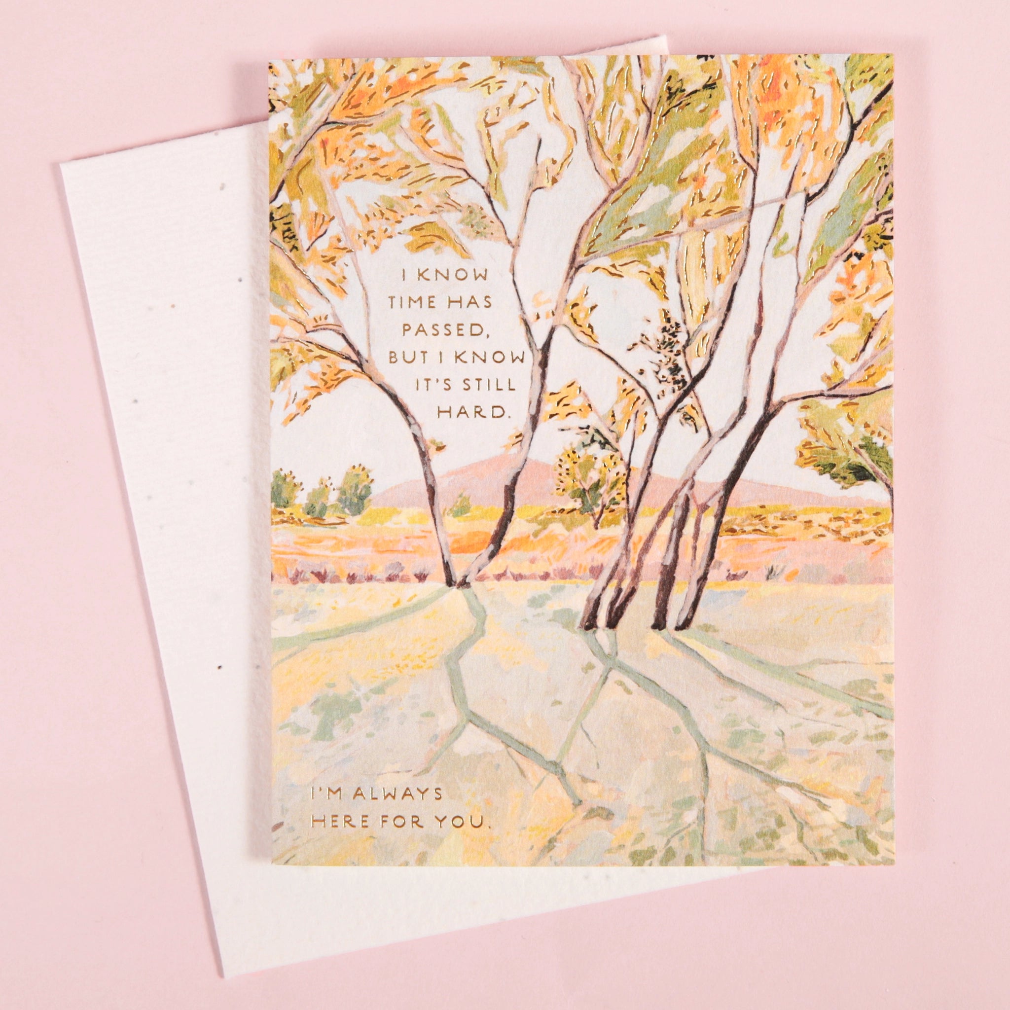 On a light pink background is a white envelope and card with a yellow and light green illustration of trees in a park with text that reads, "I know time has passed, but I know it's still hard. I'm always here for you".