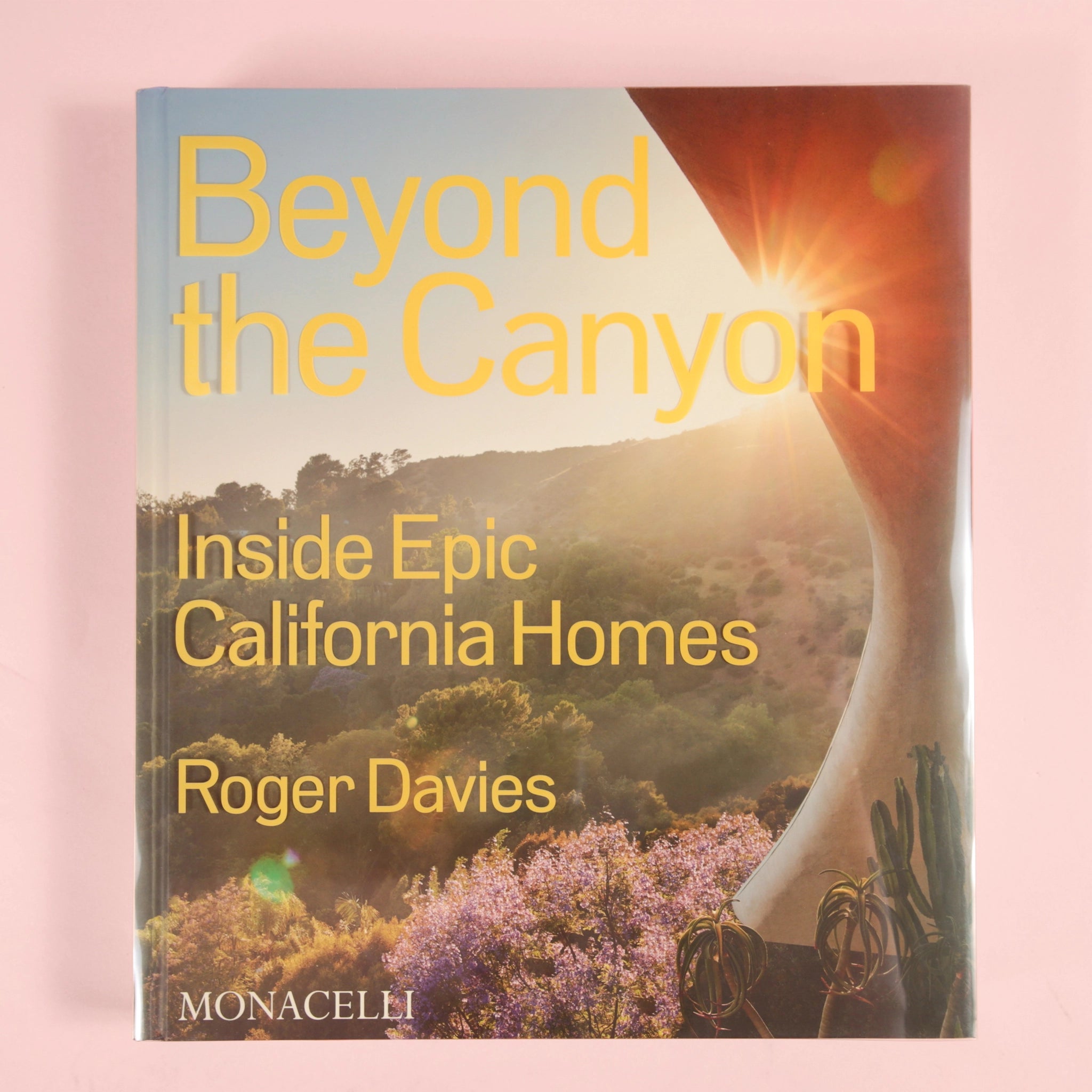 On a light pink background is a book cover with a photograph of a canyon and the title that reads, "Beyond the Canyon Inside Epic California Homes" in yellow letters. 