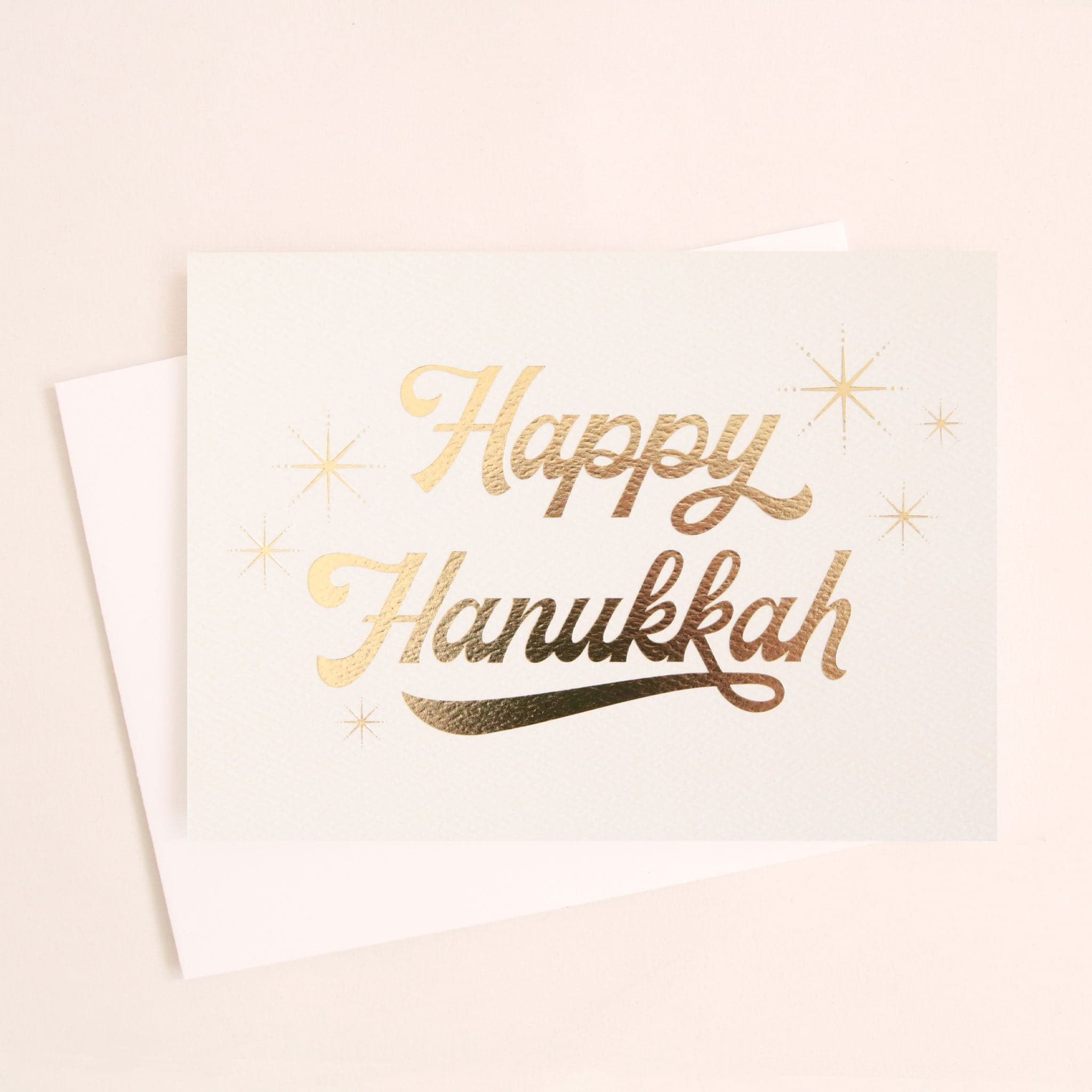 ivory greeting card with gold foil cursive text that reads happy Hanukkah surrounded by six gold foil starbursts