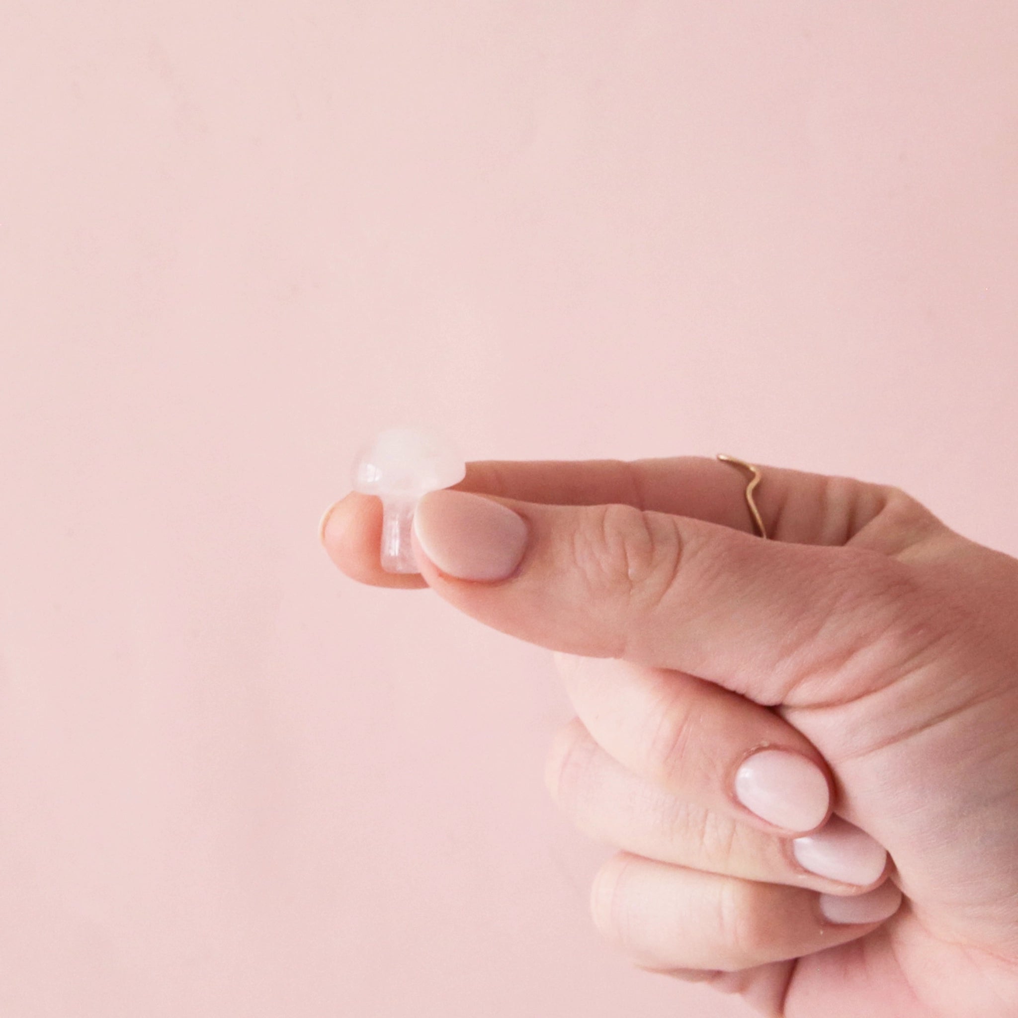 On a light pink background is a model holding a tiny rose quartz crystal in the shape of a mushroom.