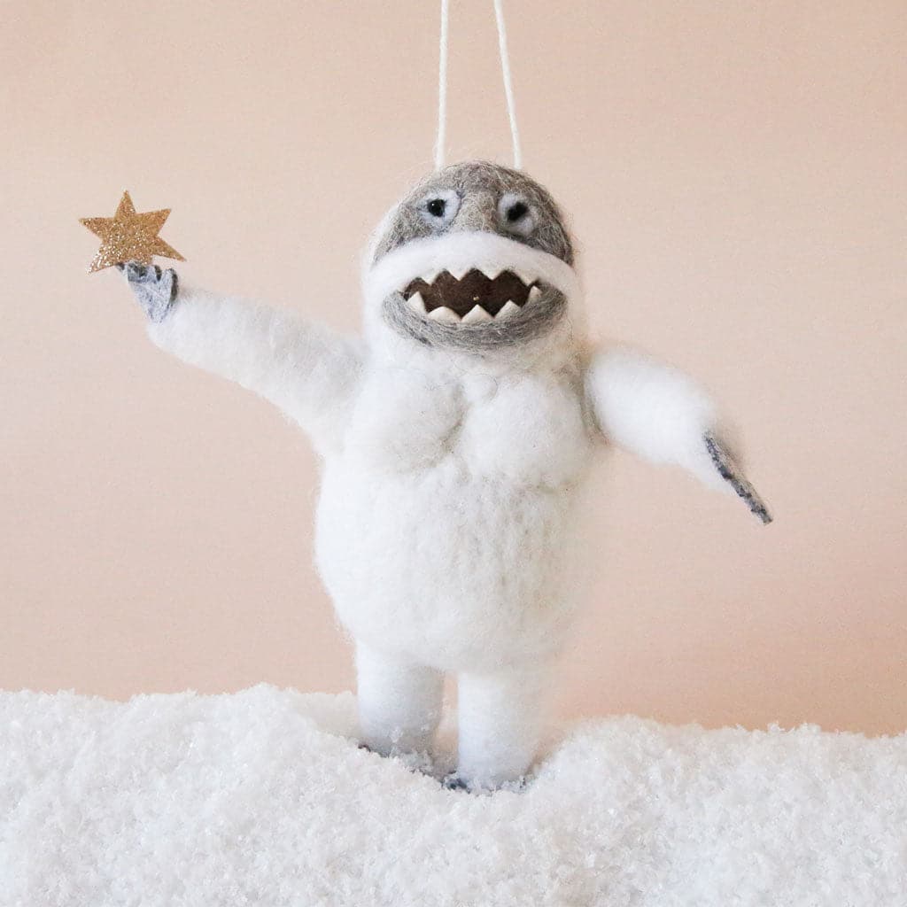 On a peach background is a white and gray abominable snowman ornament made of felt and holding a star.