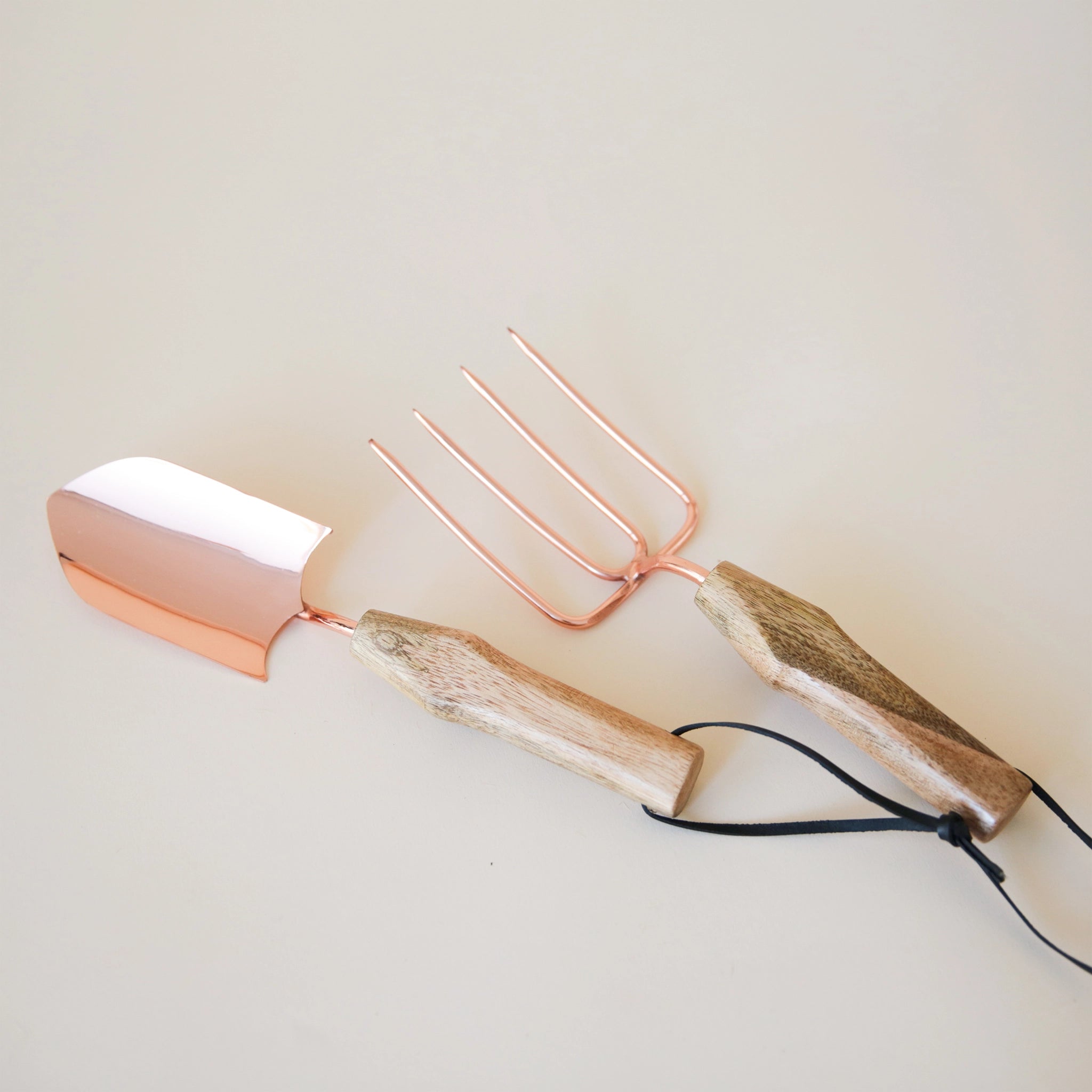 On a cream background is a set of gardening tools that have a rose gold metal, mango wood handles and black leather loops on the end. The set includes a hand shovel and a gardening fork. 
