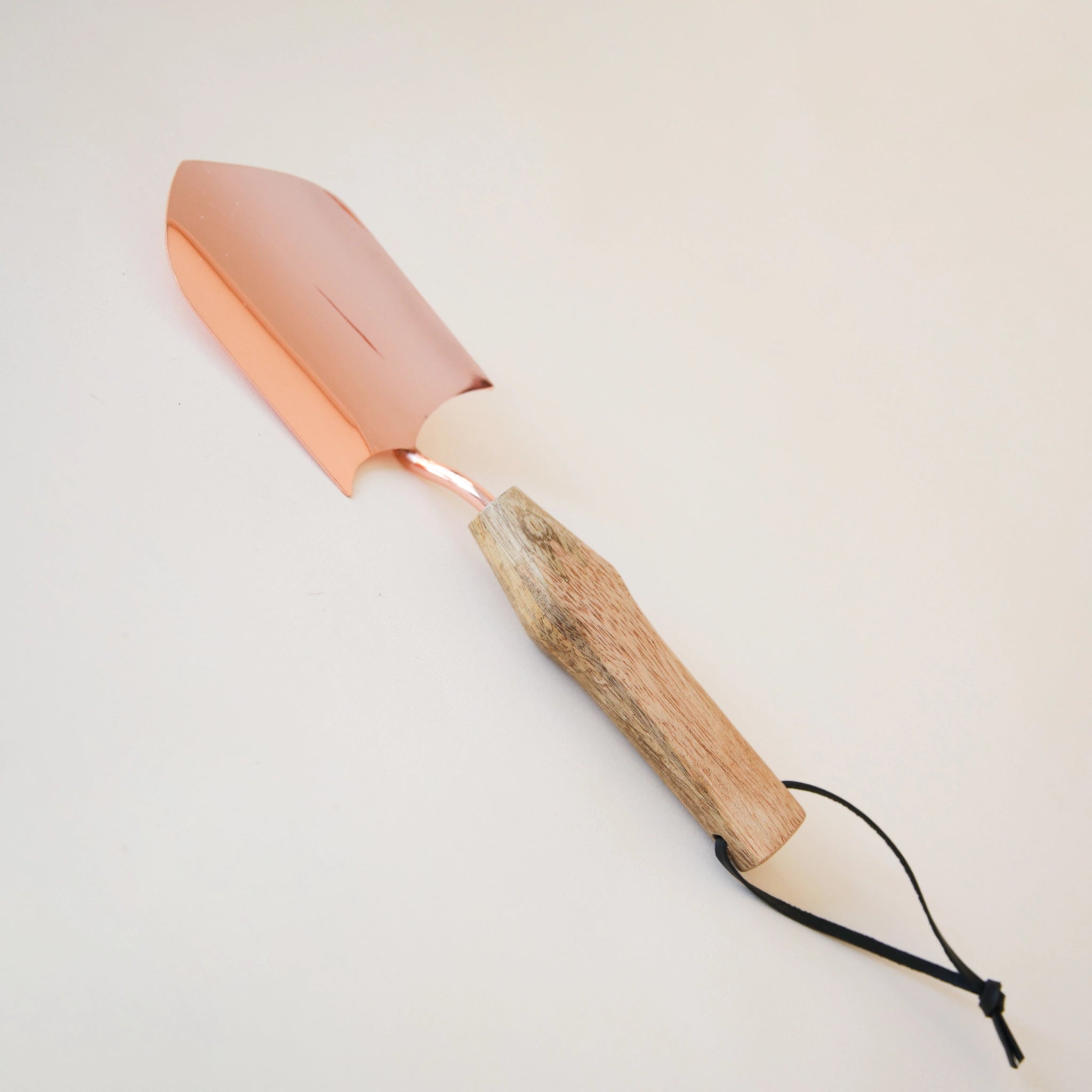 On a cream background is a set of gardening tools that have a rose gold metal, mango wood handles and black leather loops on the end. The set includes a hand shovel and a gardening fork.