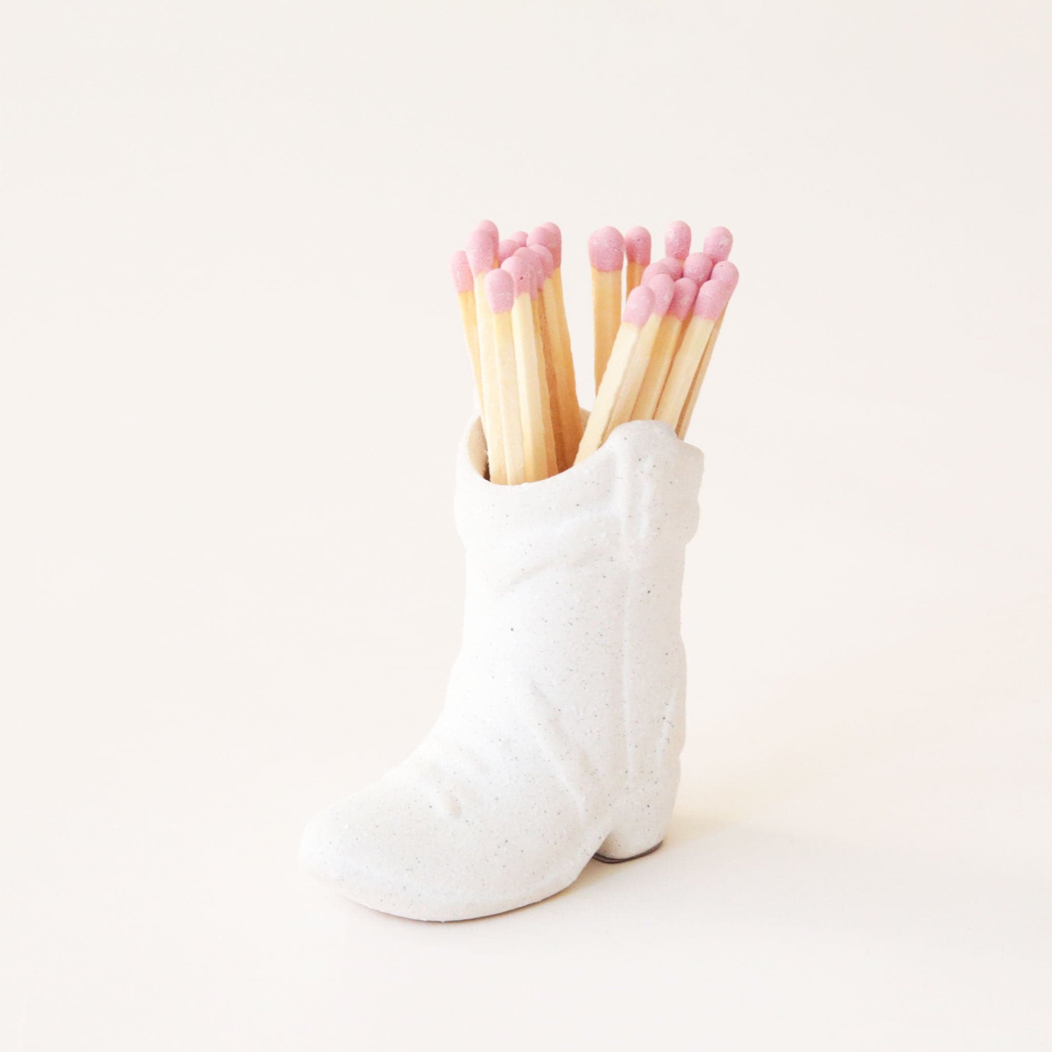 On a white background is a white cowgirl boot shaped match holder with pink matches inside.