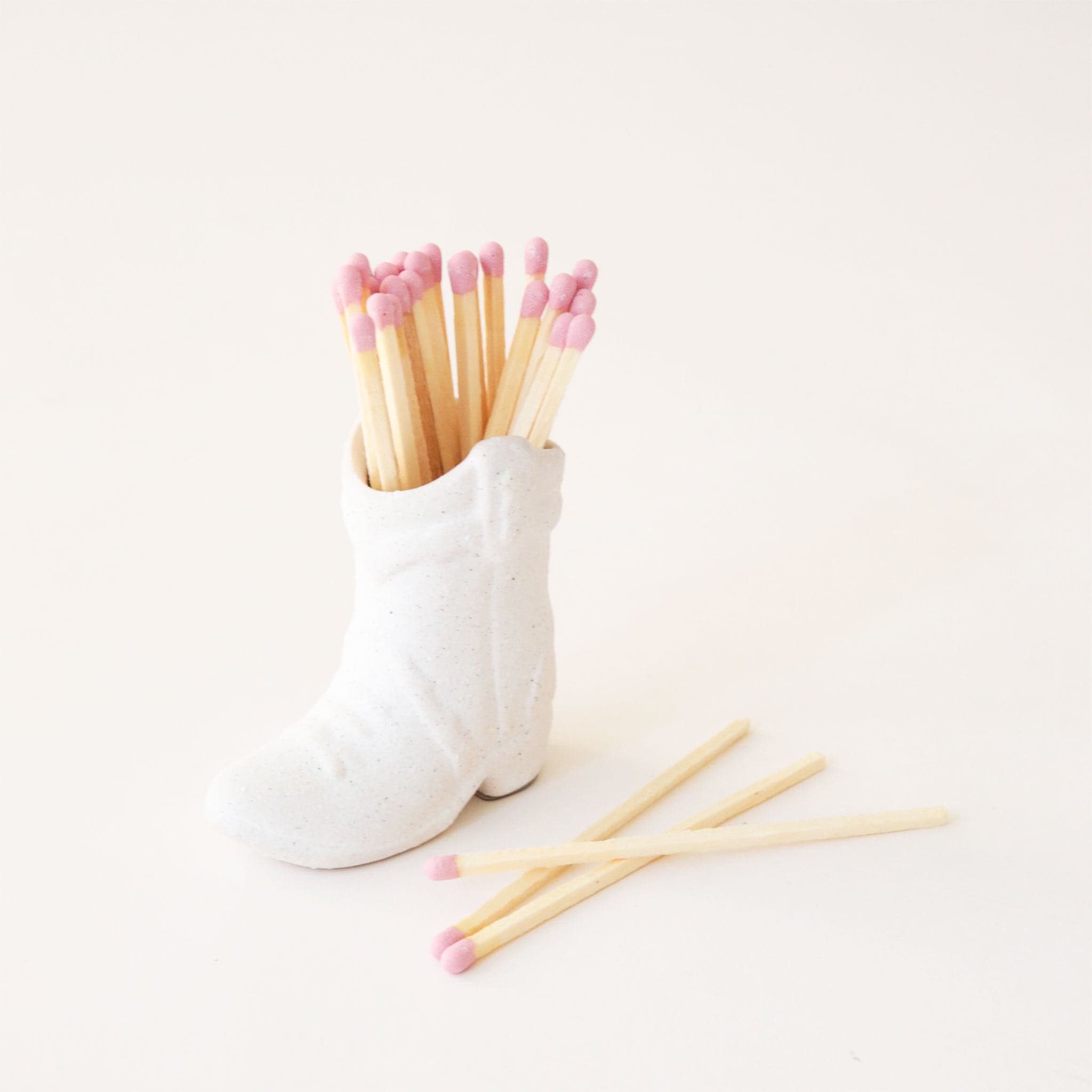 On a white background is a white cowgirl boot shaped match holder with pink matches inside. 