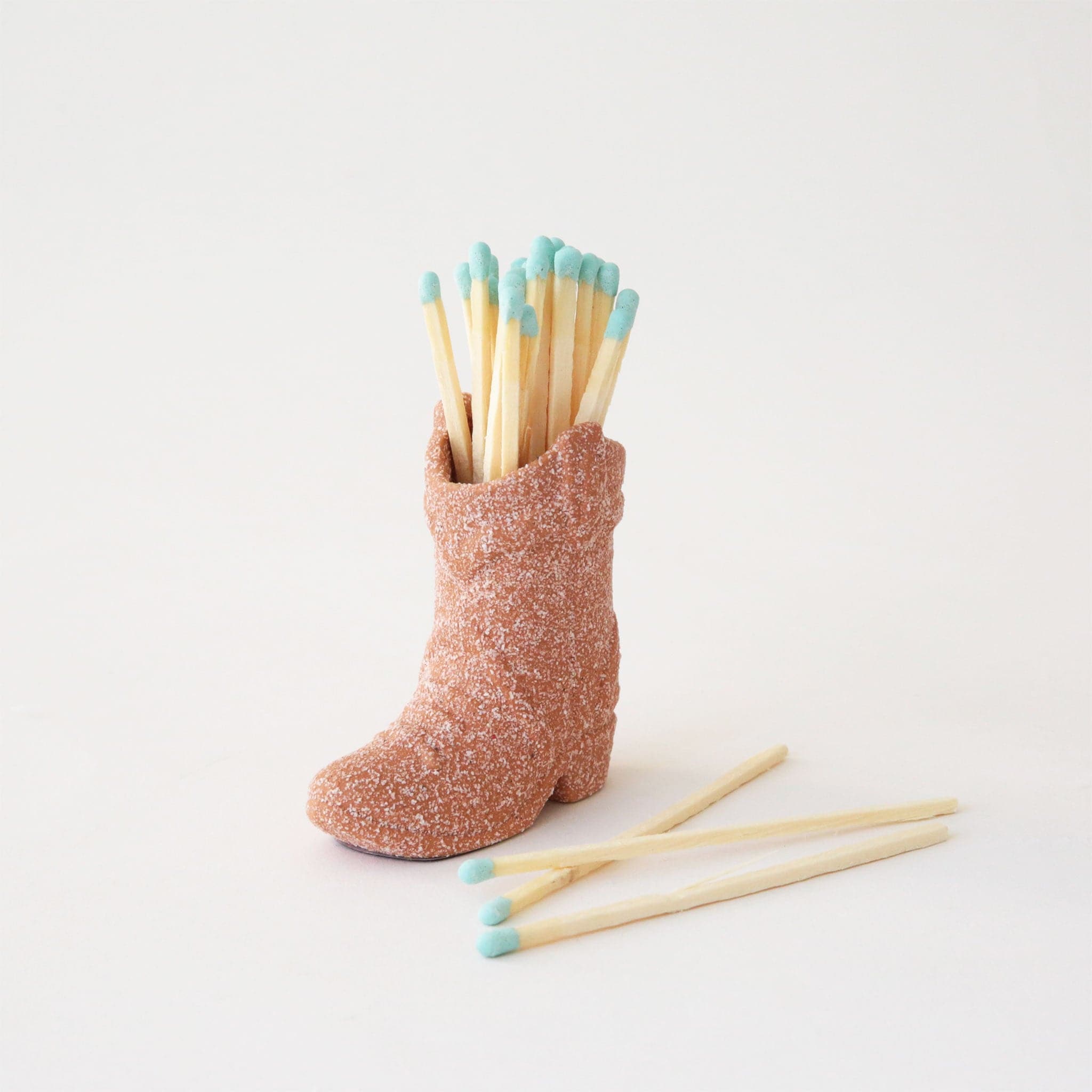 Terracotta ceramic boot match holder filled with a bundle of baby blue dipped matches. Three matches lay loose in front.