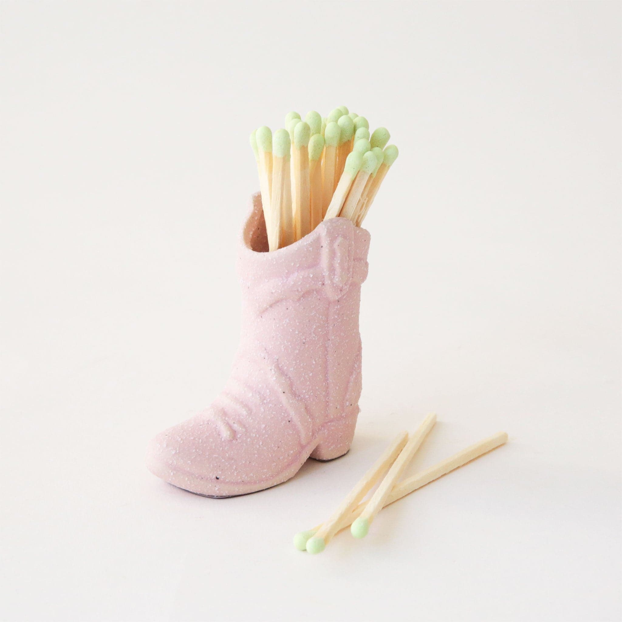 On a white background is a pink ceramic cowboy boot shaped match holder, photographed here with matches. 