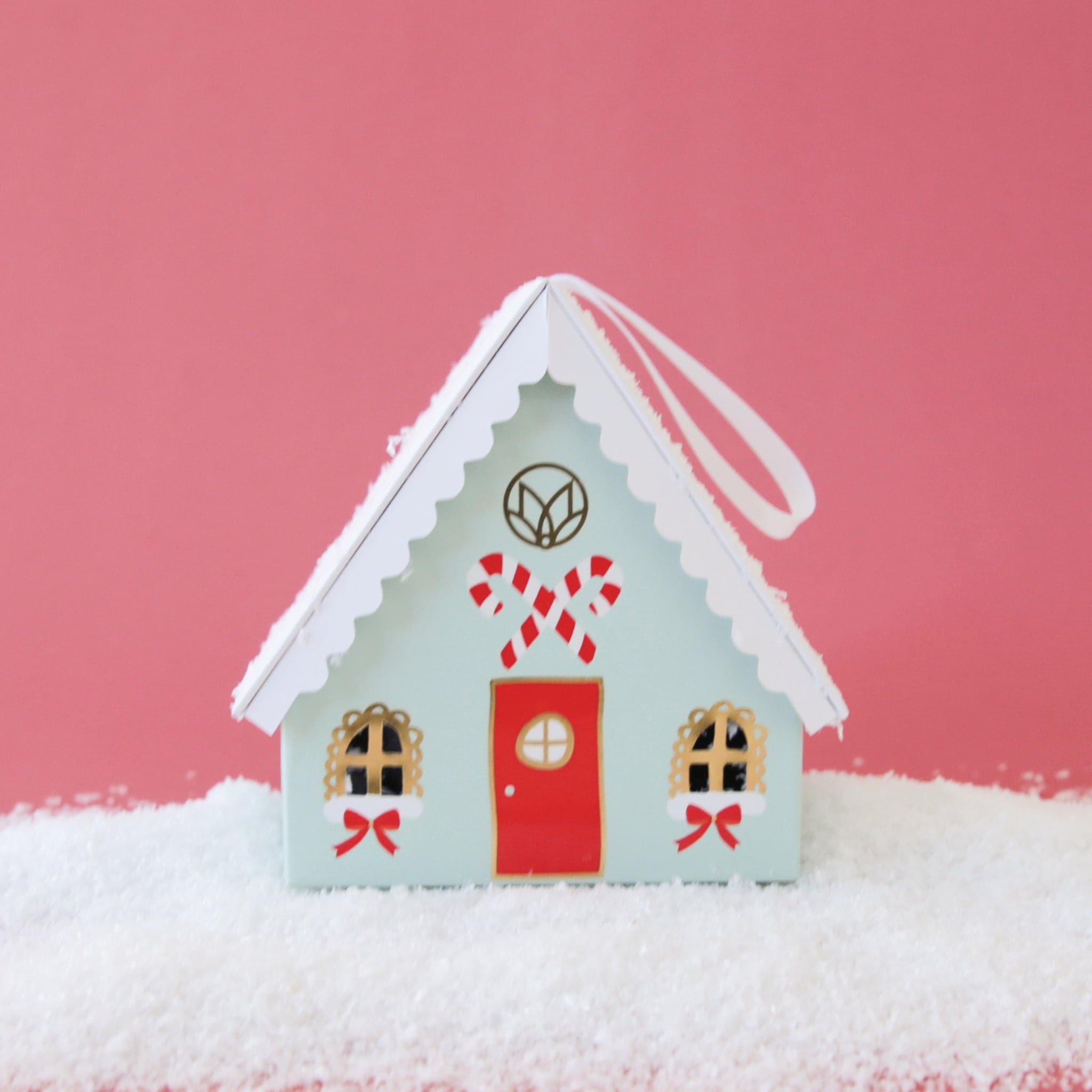 A blue holiday cottage that is home to a blue bath bomb. The cardboard cottage comes with a ribbon loop at the top that makes it double as an ornament.