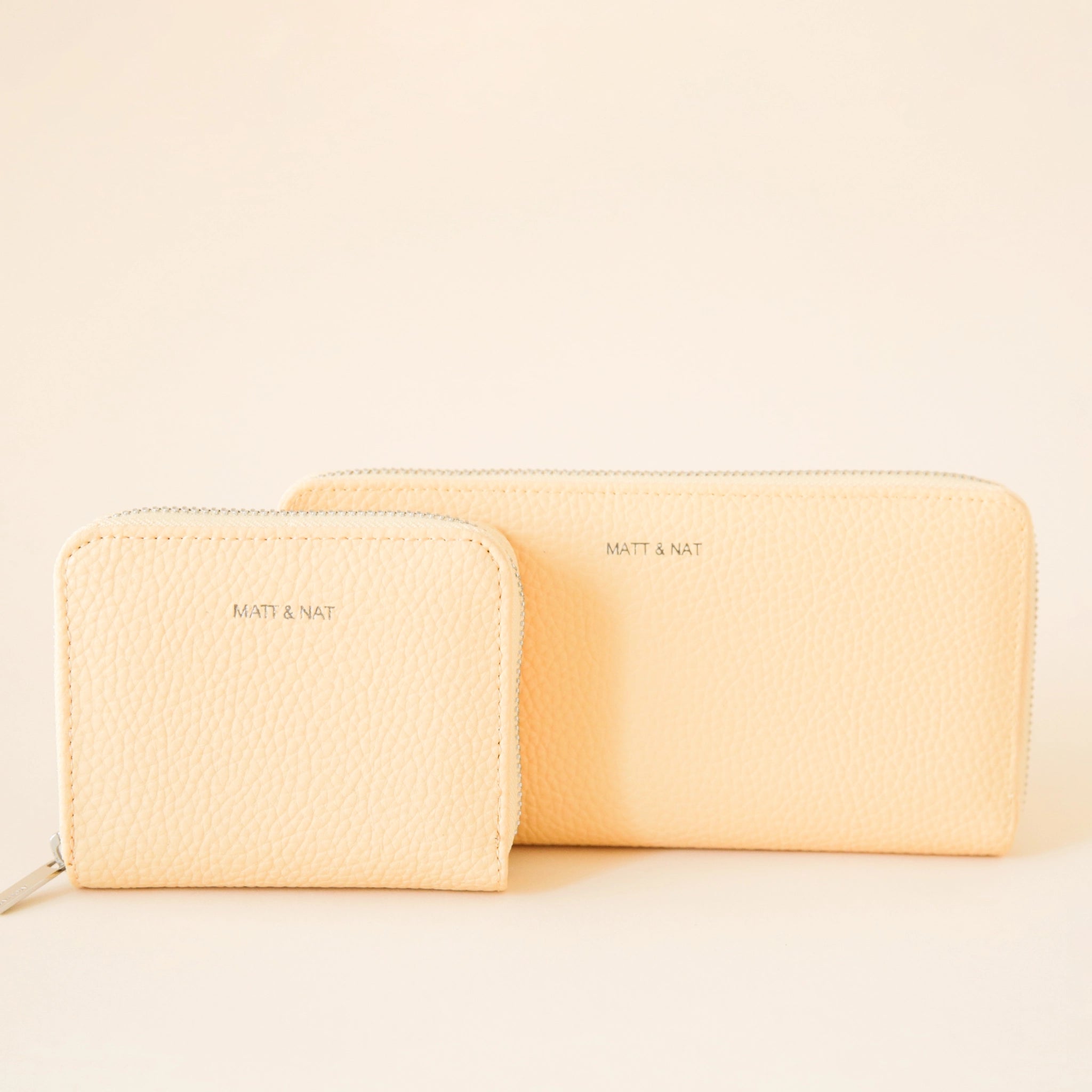 On a cream background is a light yellow zip wallet with silver detailing and "Matt & Nat" written tiny on the front of the wallet, photographed with a smaller square shaped wallet. 