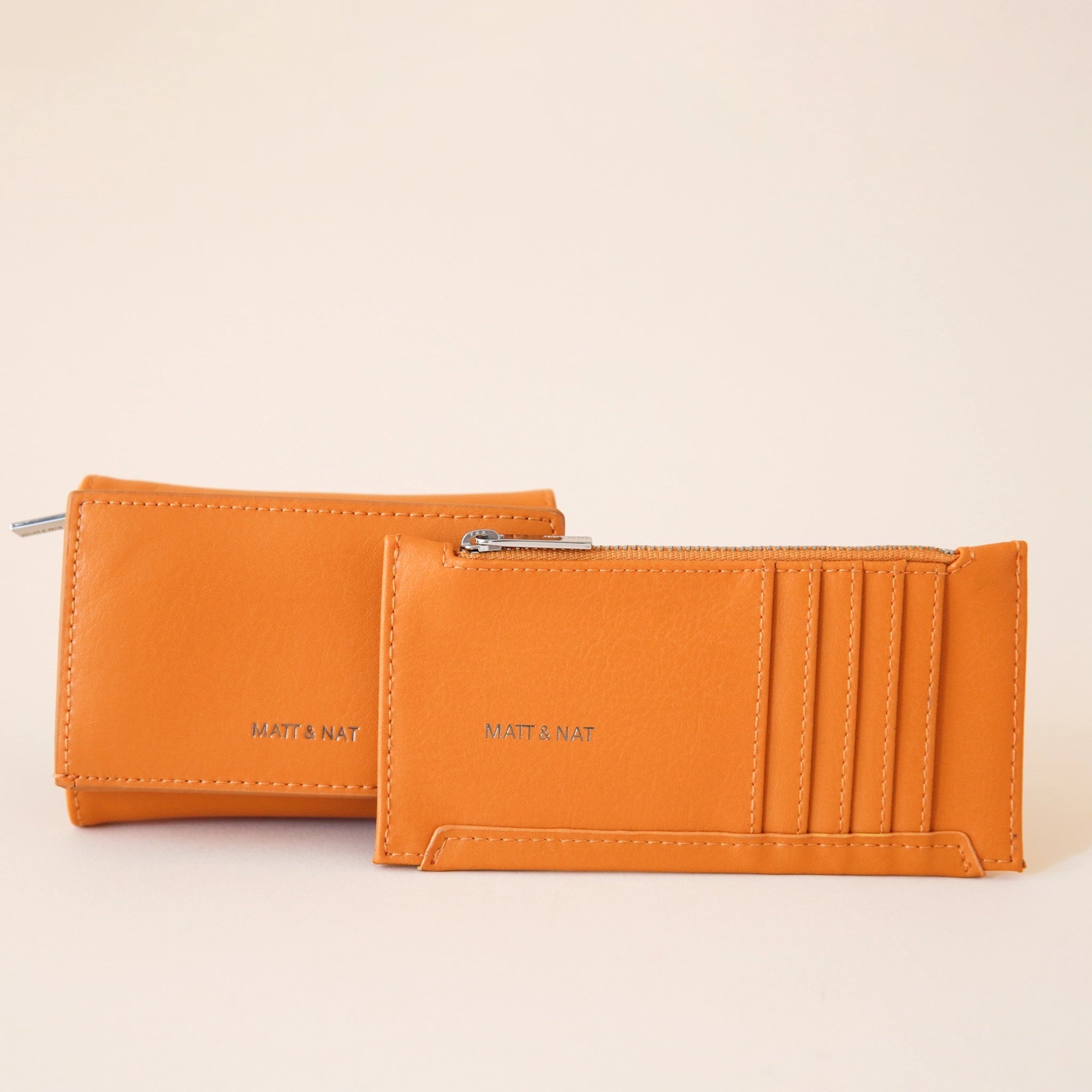 On a cream background is a bright orange colored squares wallet with a folding detail and tiny text on the bottom of the front that reads, "Matt & Nat" photographed next to a card holder that we also sell on our website. 