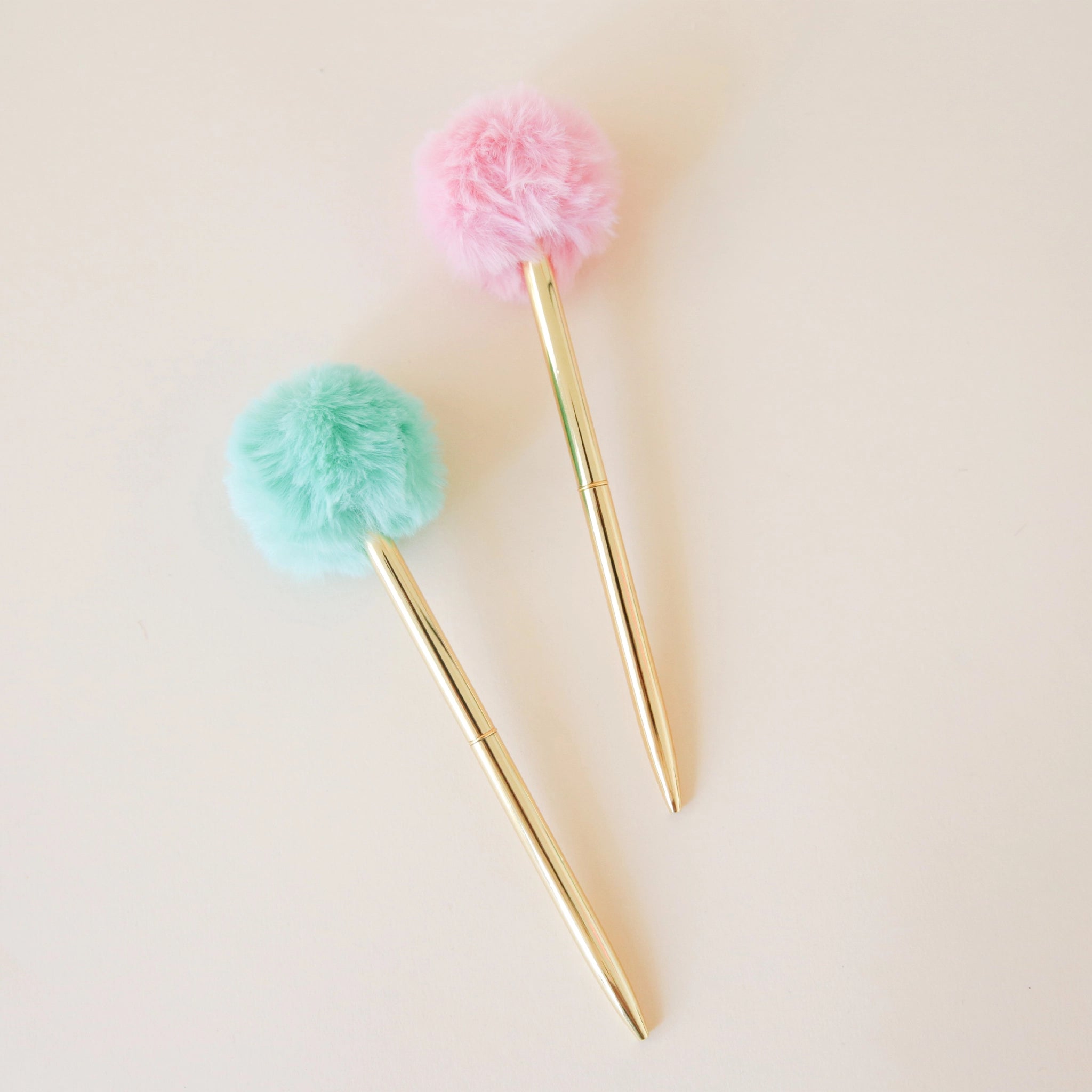 two thin gold pens. one with a fluffy mint green pom pom at the end and the other with a pink pom pom