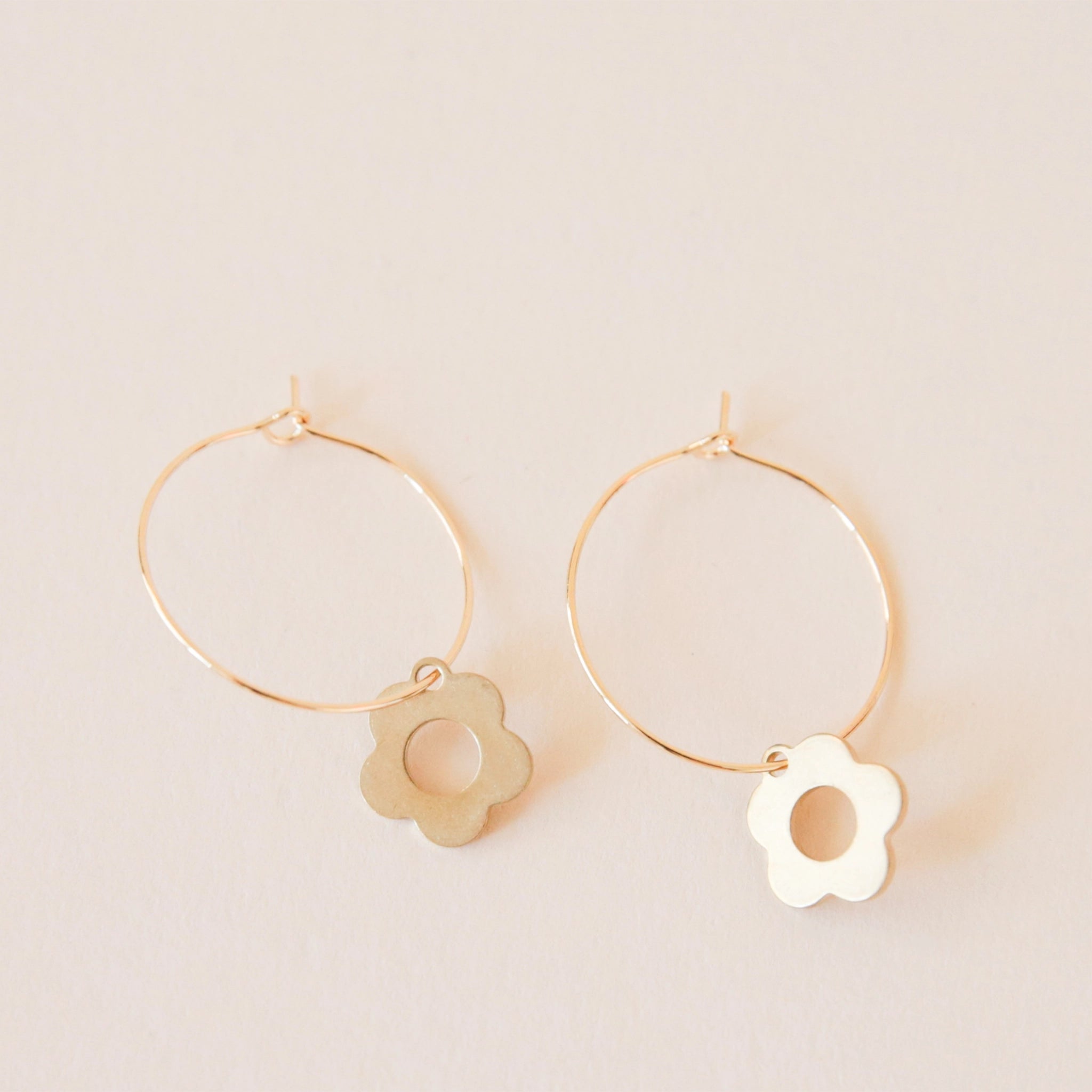 On a cream background is a dainty gold hoop earring with a smaller daisy charm dangling from the bottom edge. 