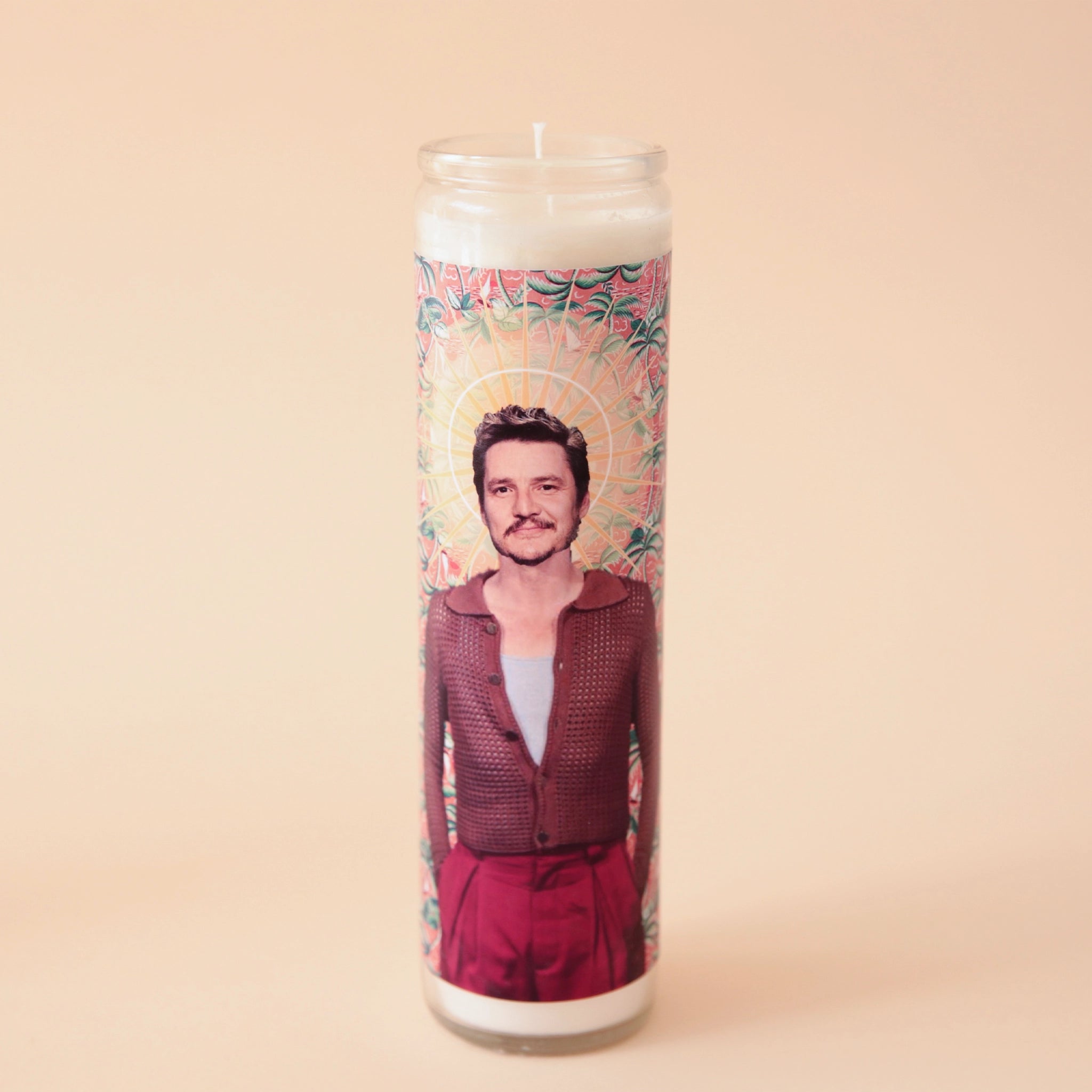 On a cream background is a tall thin prayer candles in a clear glass jar with a label around it that features a photograph of Pedro Pascal on the front in a red outfit. 
