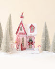 a pink and white holiday house surrounded by silver bristle tress on a ground of snow