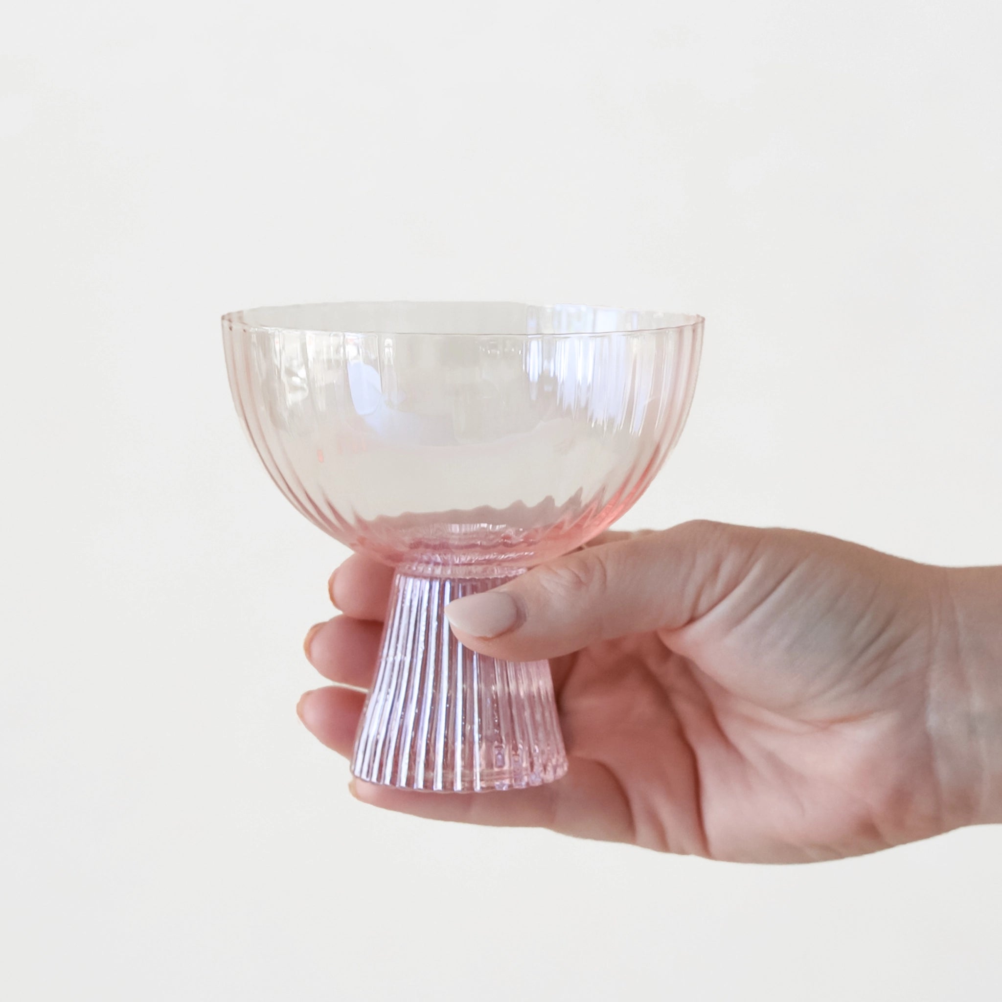 On a white background is a ribbed light pink coupe glass being held by a models hand. 