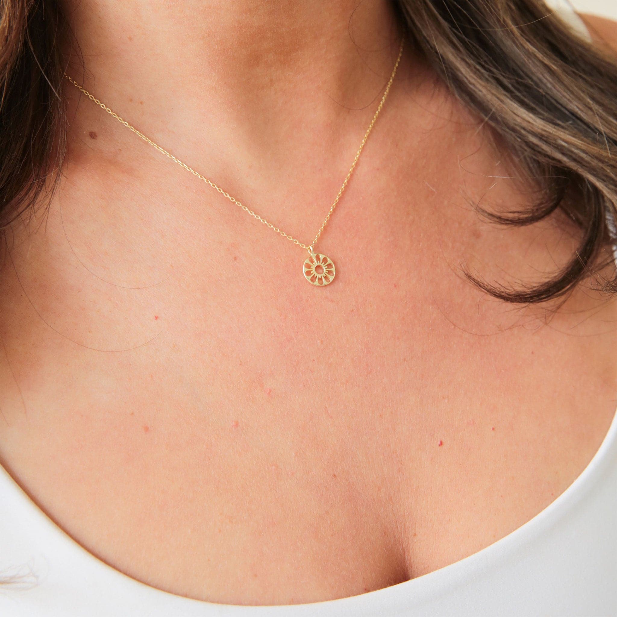 A dainty gold chain necklace with a gold circle pendant in the center with a cut out of a daisy flower, photographed being worn on a model.