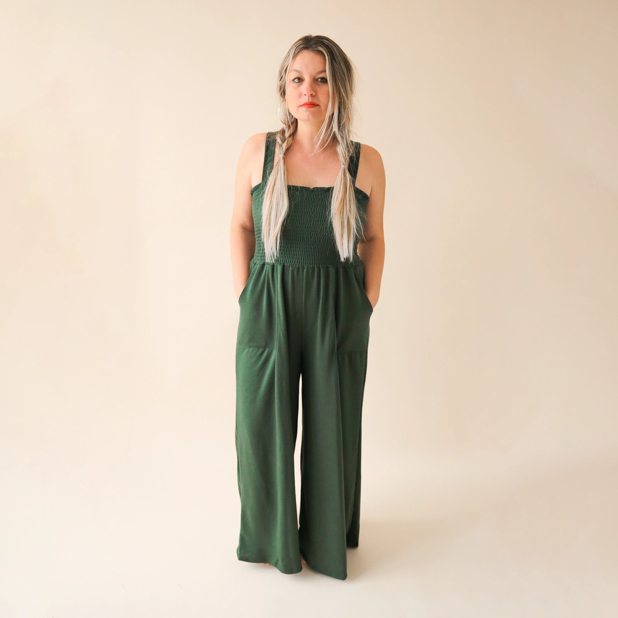 On a cream background is a model wearing a hunter green wide leg jumpsuit with large pockets and 1.5" shoulder straps.