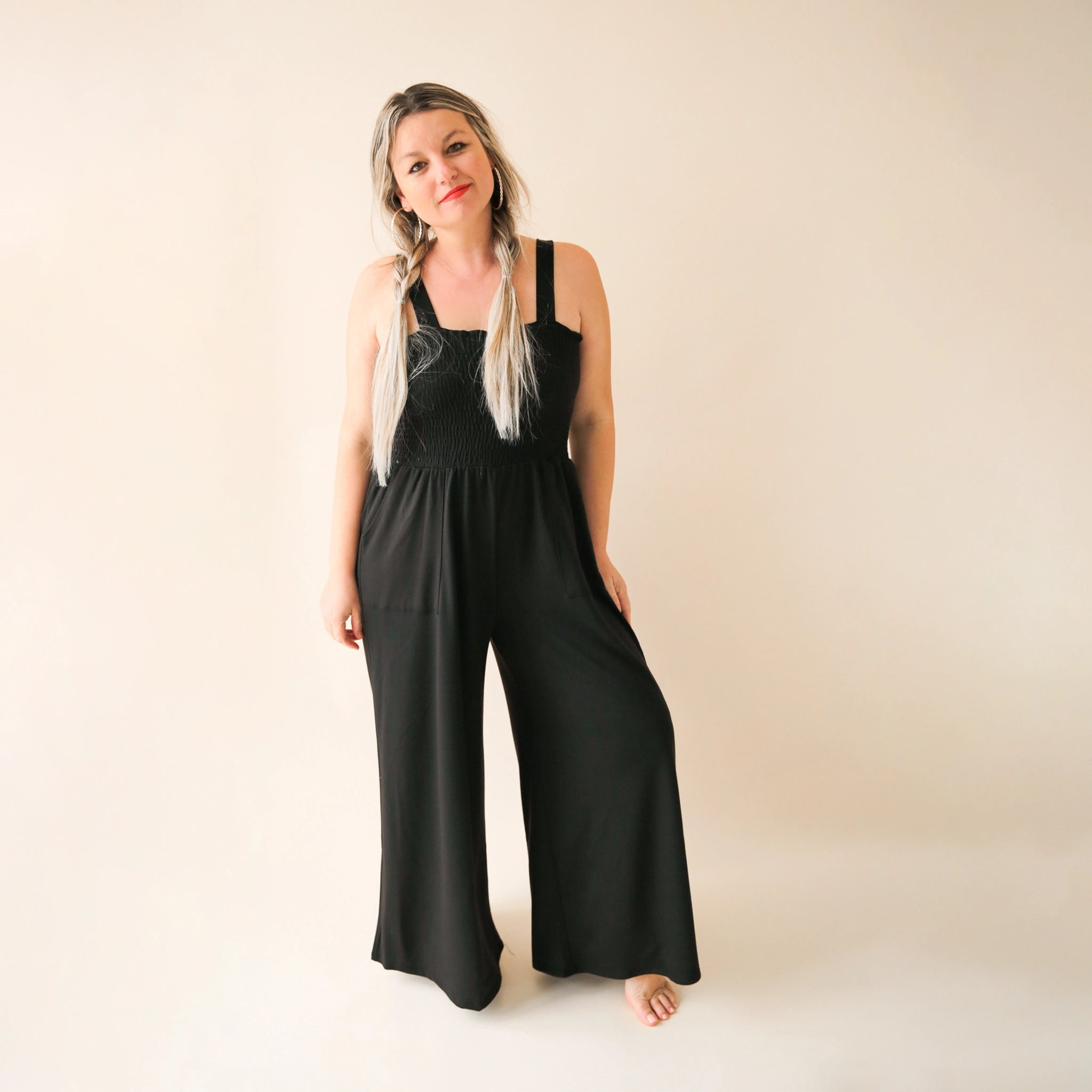 On a cream background is a model wearing a black wide leg jumpsuit with large pockets and 1.5" shoulder straps. 