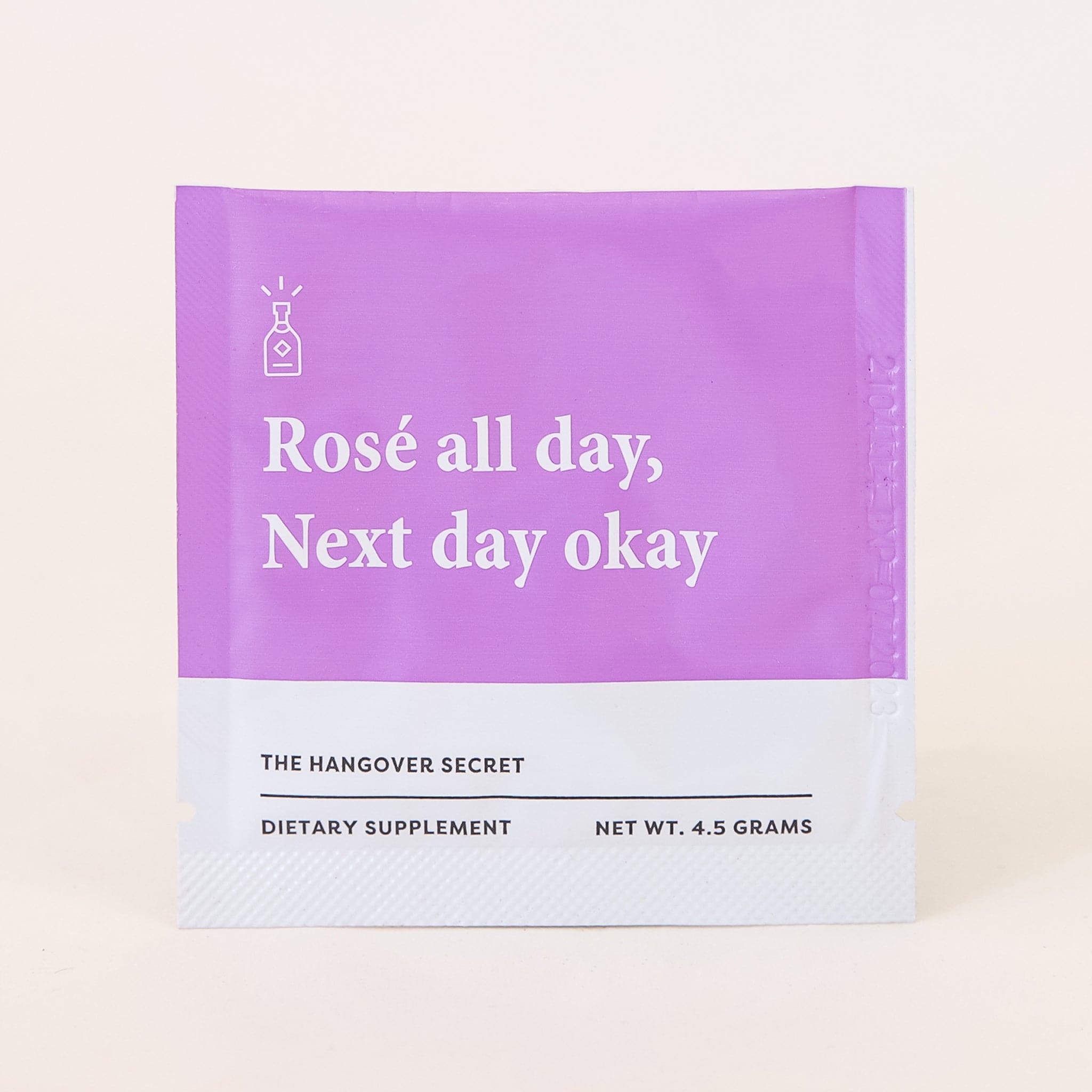 A small packet filled with a dietary supplement. The packaging's top half is a bright purple and the bottom half is white. There is text across the front that reads, "Rosé all day, Next day okay" as well as "The Hangover Secret | Dietary Supplement".