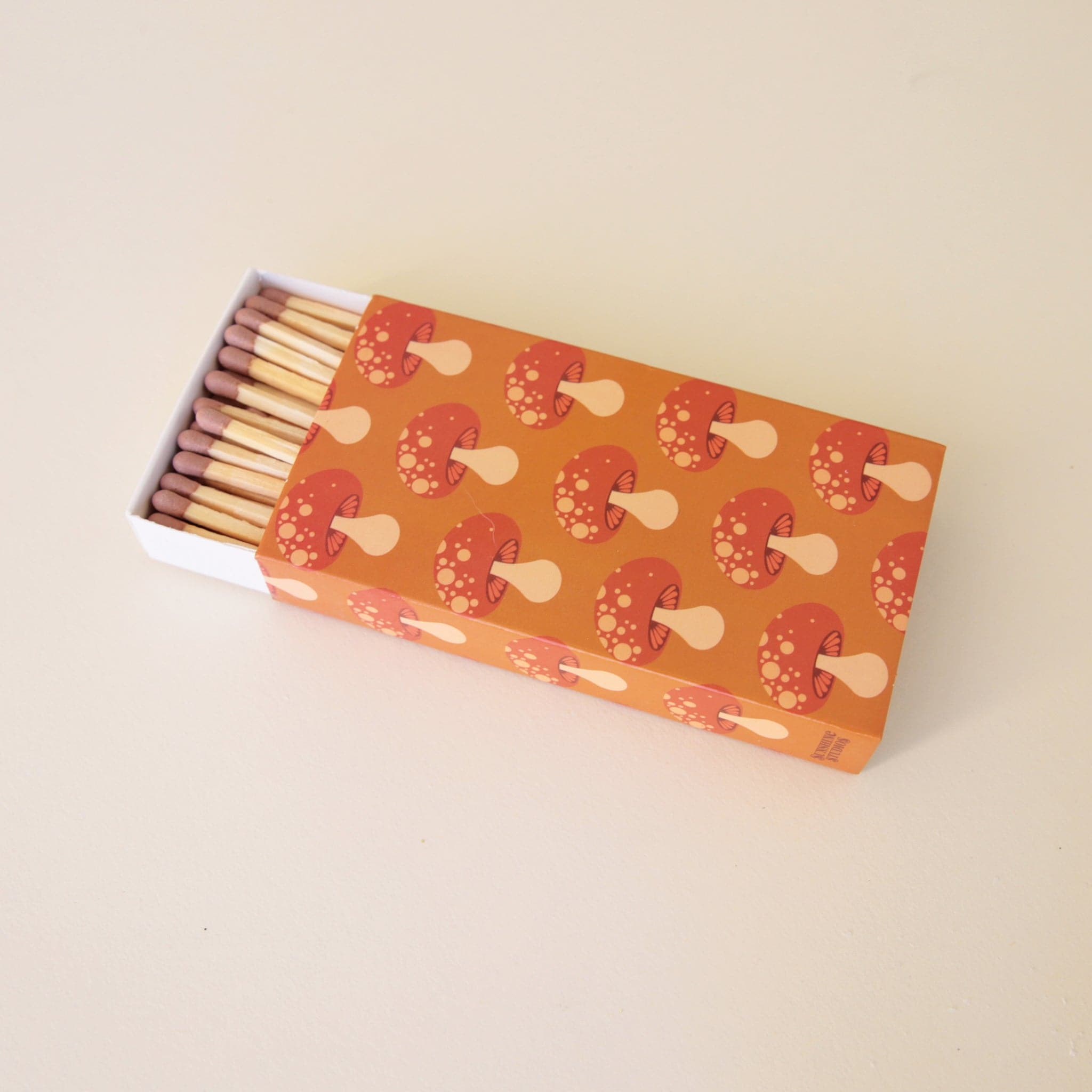 A rectangle box of matches with a repeating red mushroom design along all edges besides the top and bottom. The matches that are inside are also photographed here. They are wooden matches the same length as the box with a red striking tip for lighting.  Edit alt text