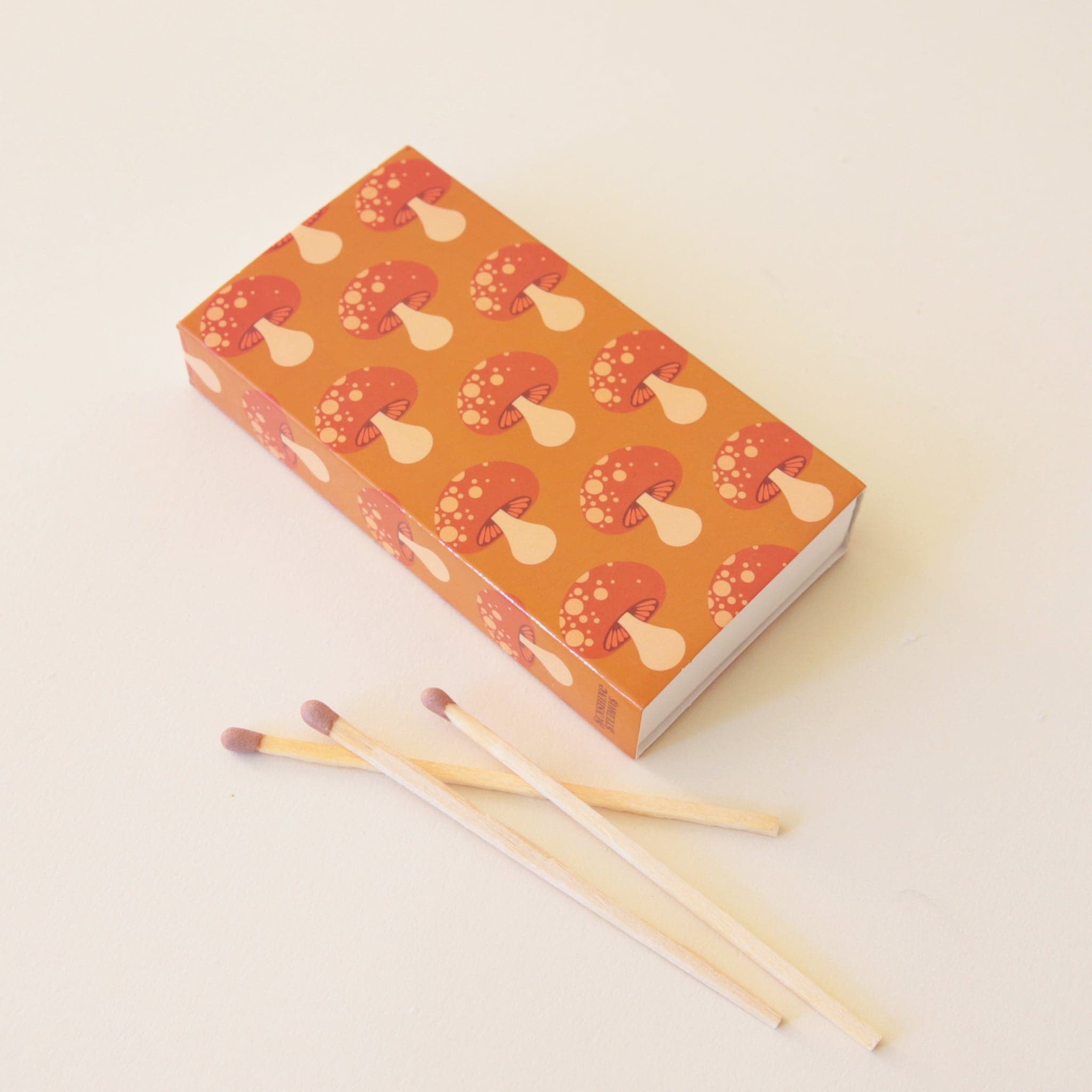 A rectangle box of matches with a repeating red mushroom design along all edges besides the top and bottom. The matches that are inside are also photographed here. They are wooden matches the same length as the box with a red striking tip for lighting.
