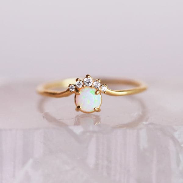 In front of a white background is a gold ring. On the front is a round white opal with five white stones on the top of the opal.