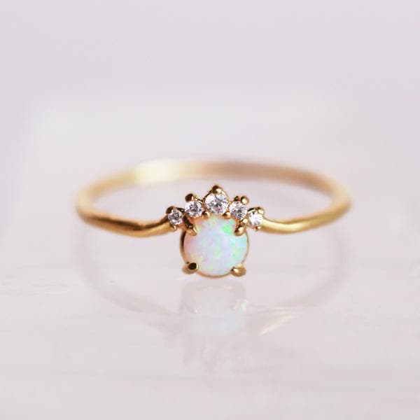 In front of a white background is a gold ring. On the front is a round white opal with five white stones on the top of the opal. 