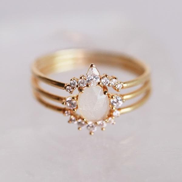 In front of a white background is a stack of three rings. Each ring has a gold band. The top ring has an upward arch in the middle. The arch has three round diamonds on each side and a tall oval diamond in the middle. The middle band has a round white crystal in the middle with a small diamond on each side. The last band has a downward arch with seven small, round diamonds on the arch.
