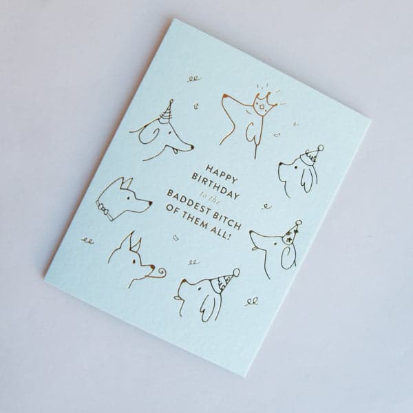Light blue illustrated greeting card with sketchy dog heads wearing party hats, in gold foil, with text "Happy Birthday to the Baddest Bitch of Them All!"