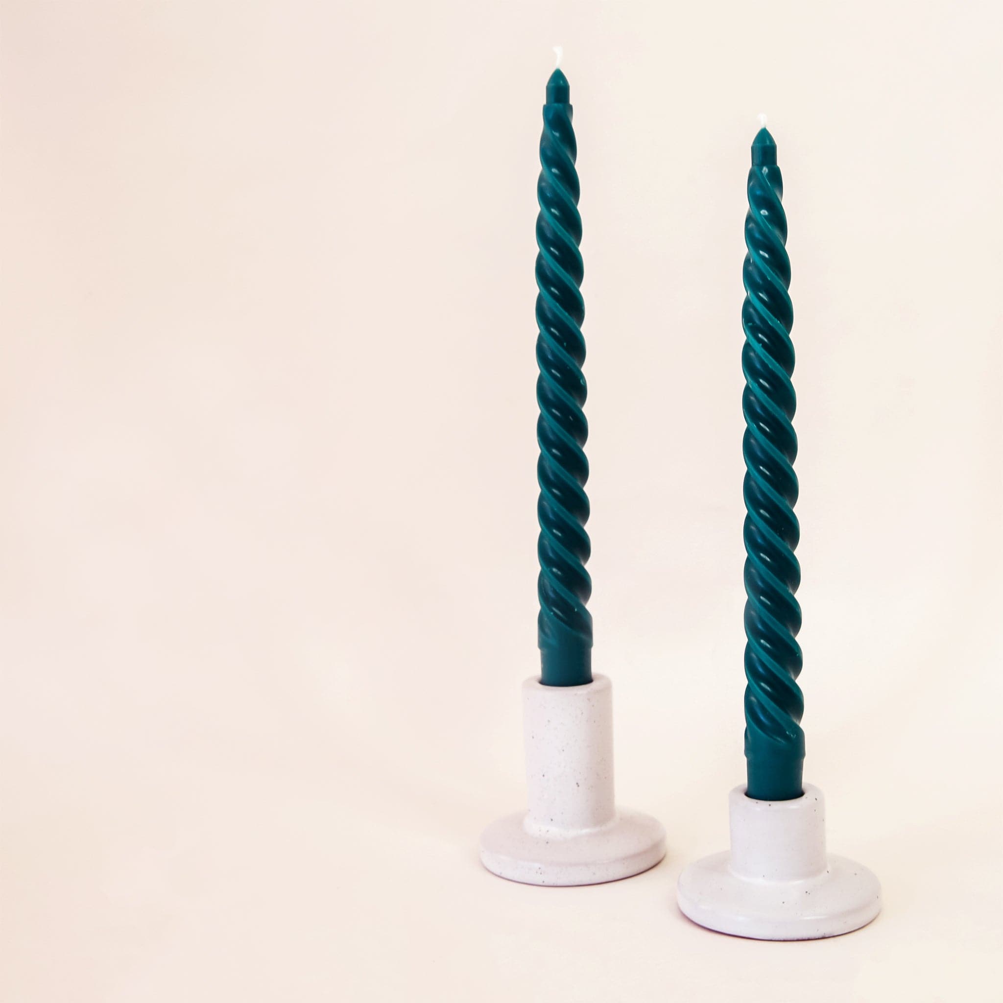 Against a white background are two gray, ceramic candle holders. The candle holder on the left has a round base and tall, cylinder holder. The holder on the left also has a round base but the cylinder holder is about half the size of the other holder. Both holders have a dark green, twisted taper candle inside. 
