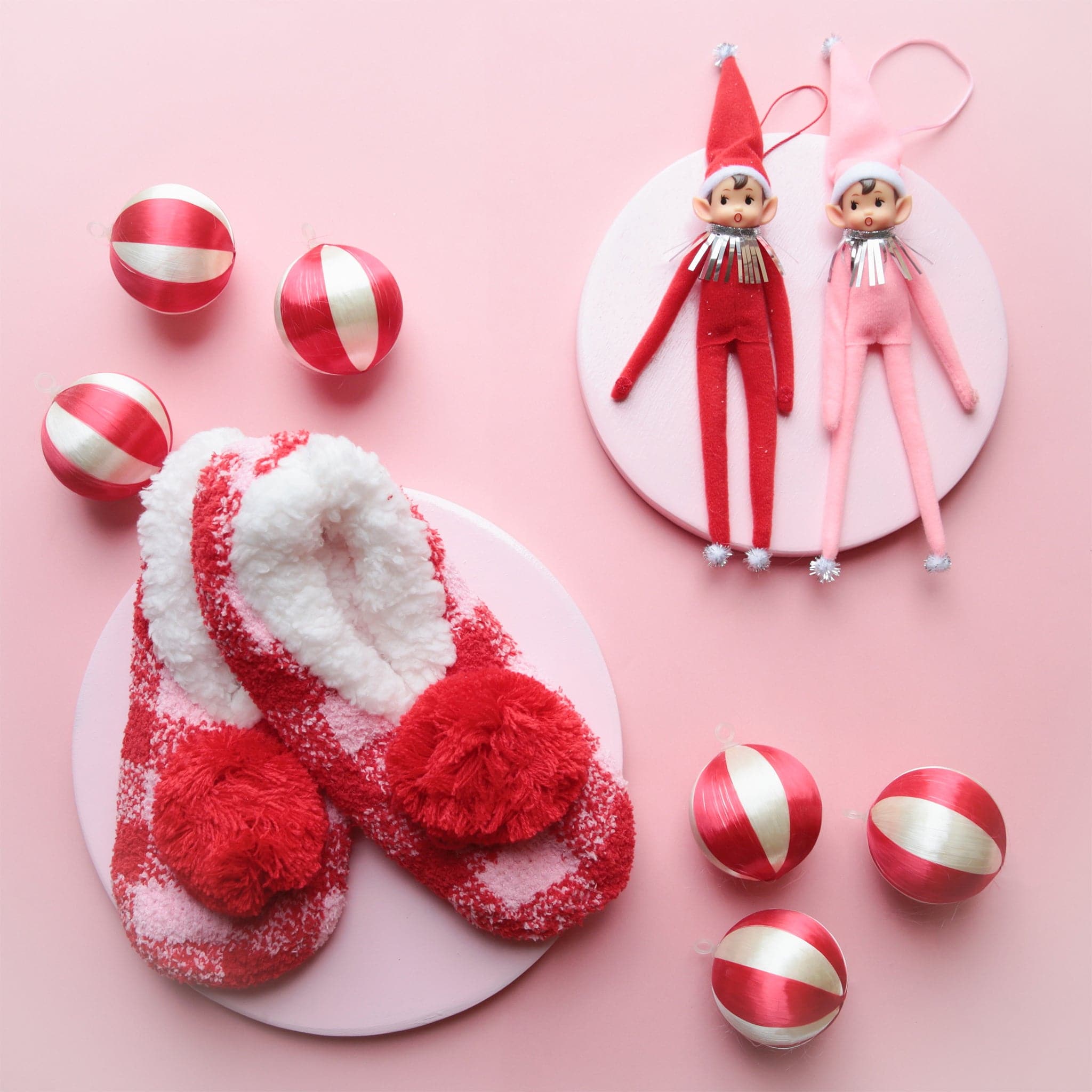 On a cream background is a elf ornament with long arms and legs with a pink suit and hat on as well as a pink string loop for hanging and photographed here next to the red version. 