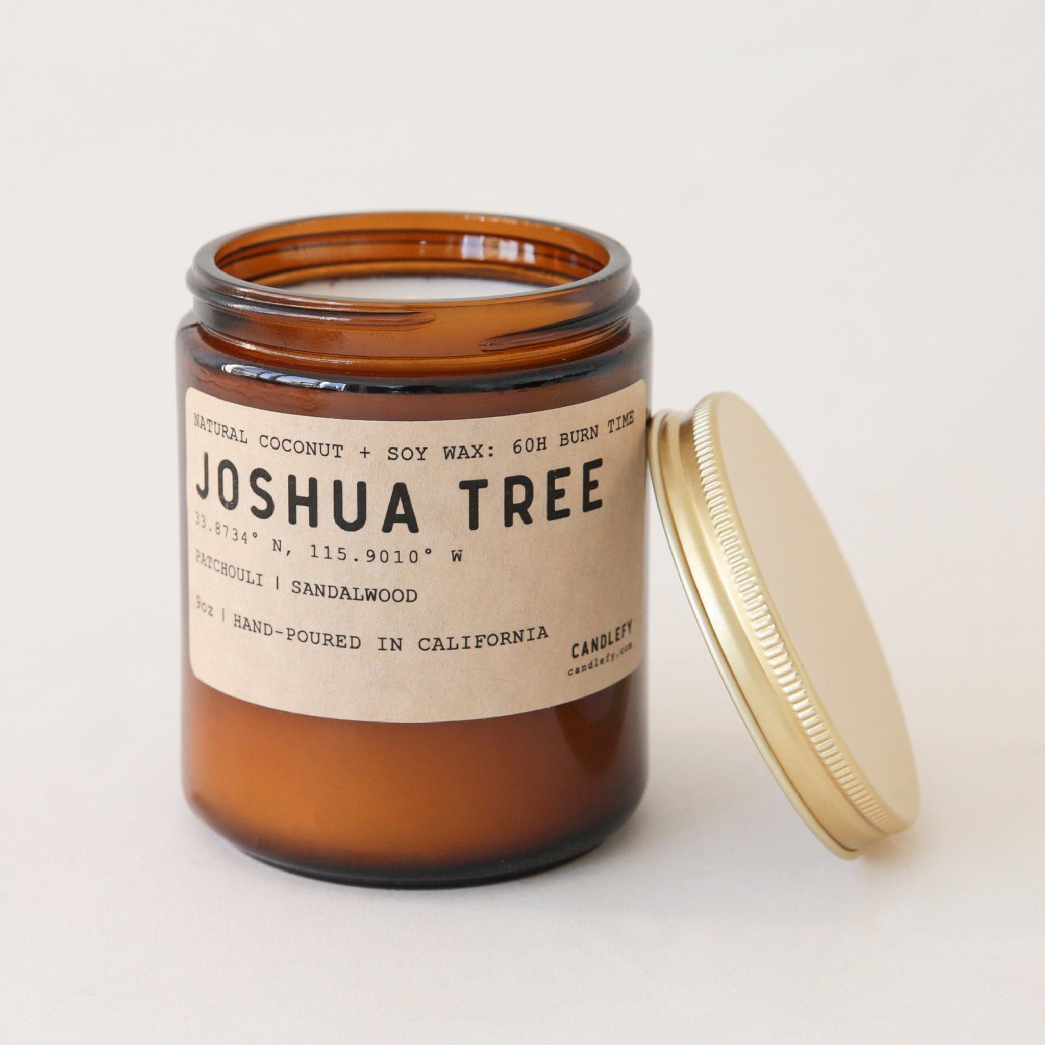 An amber colored glass candle jar with a brown label on the front that reads, "Joshua Tree Patchouli | Sandalwood" in black letter along with a single wick candle inside the jar and a gold screw on lid. 