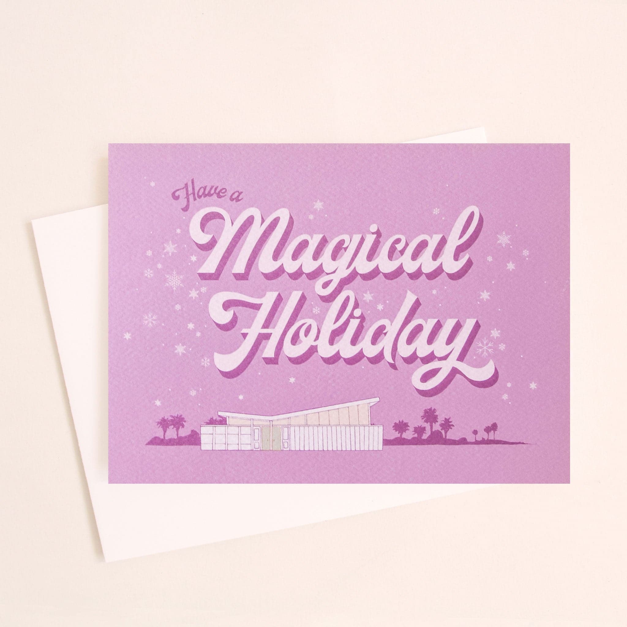 Lilac holiday card that reads &#39;have a a magical holiday&#39; in cursive lettering. Around the text is delicate white snowflakes. Below is a coastal scene of a beach house and palm trees. 