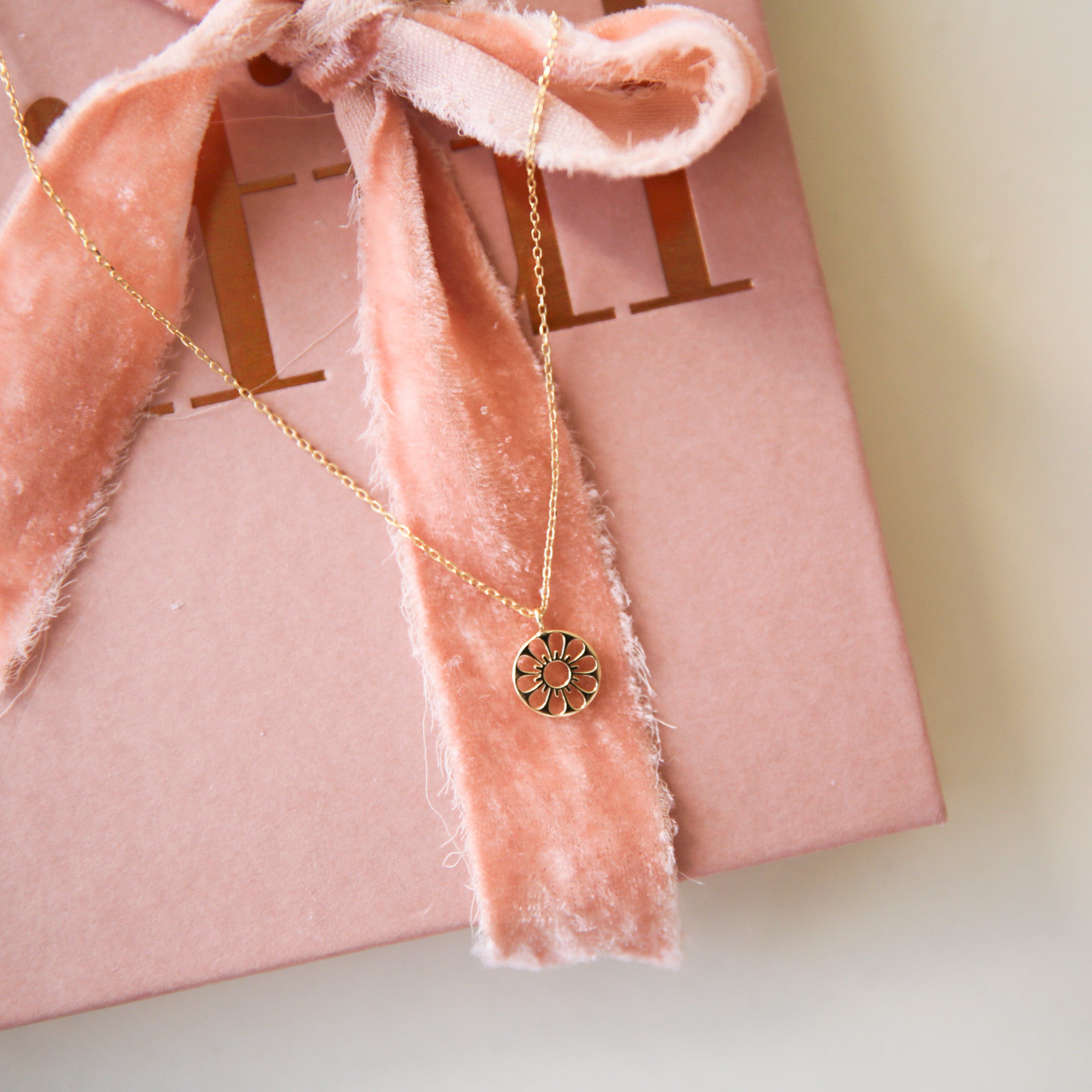 A dainty gold chain necklace with a gold circle pendant in the center with a cut out of a daisy flower, photographed on a pink book with a textured pink ribbon.
