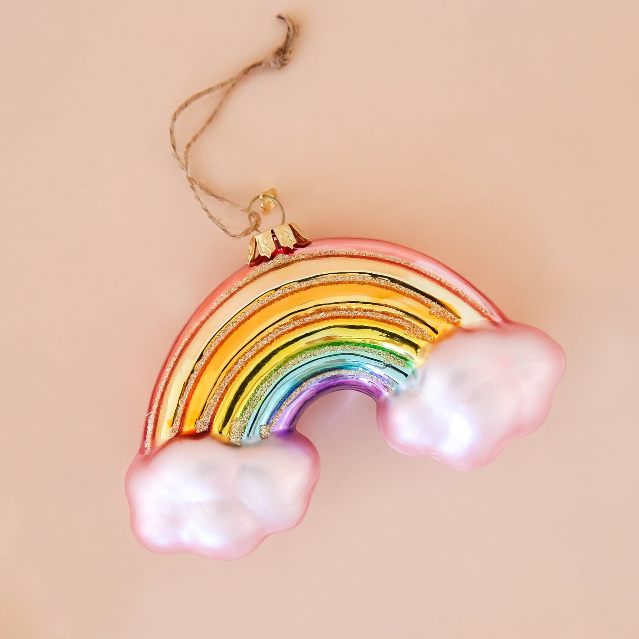 In front of a peachy background is a glass rainbow ornament. There is a light pink cloud on each end of the rainbow. The colors of the rainbow are red, orange, yellow, green, blue and purple. There is gold glitter in between each color. At the top of the ornament is a gold hook with brown twine string. 